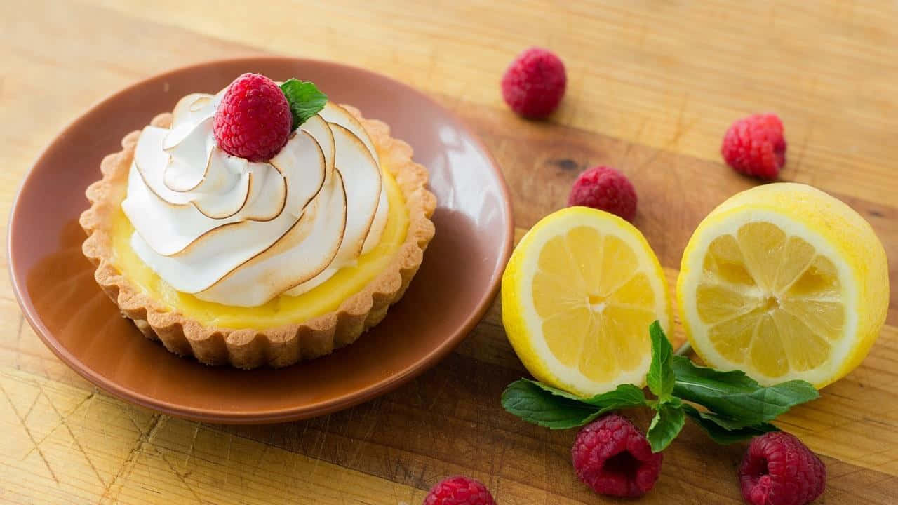 Pastry 桌布 壁紙 — 一個檸檬蛋白撻，上面放有覆盆子和薄荷，放在木板上，旁邊有切片檸檬和散落的覆盆子。
