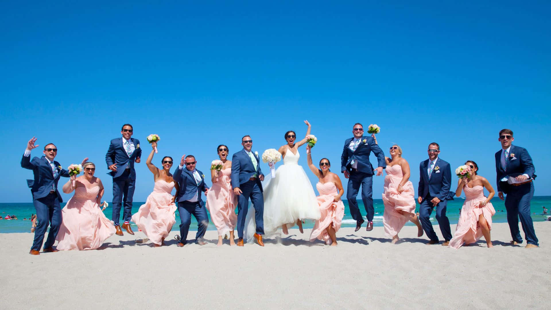 Beach Party 桌布 壁紙 — 一個歡樂的婚禮派對在陽光明媚的海灘上充滿活力地跳躍，背景是碧綠的海洋和清澈的藍天。