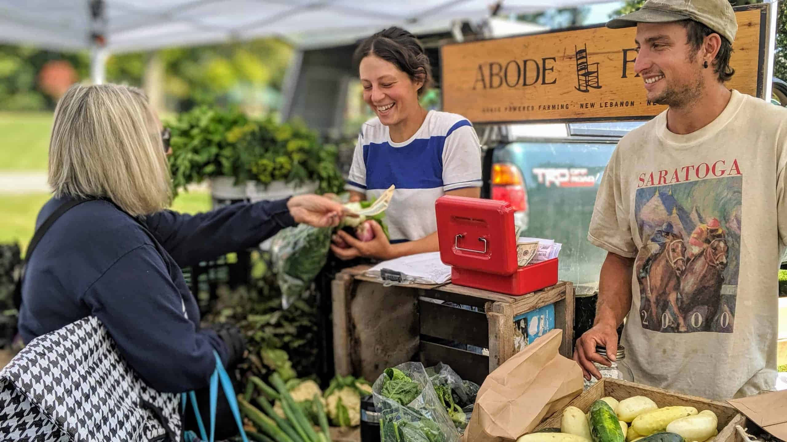 Farmers Market 桌布 壁紙 — 一個歡樂的農夫市集攤位，顧客在白色遮陽篷下從微笑的攤販手中接過新鮮農產品。