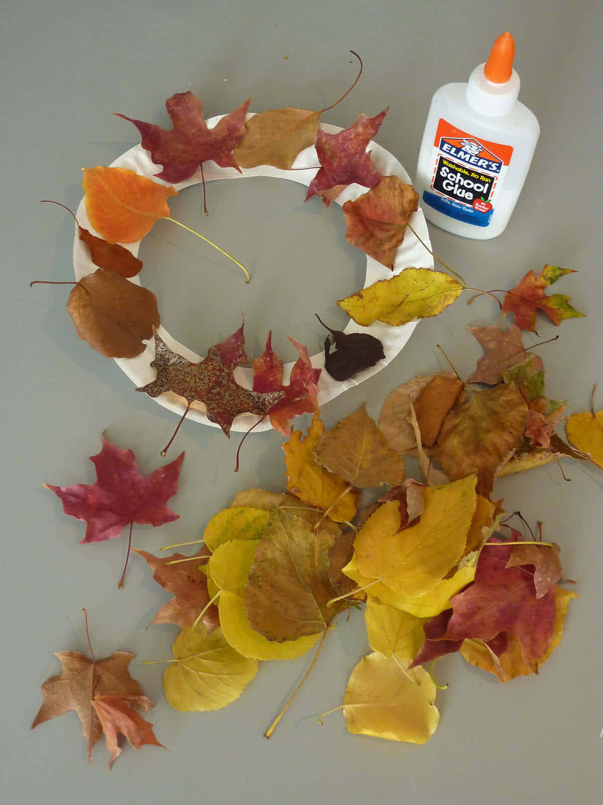 Fall Crafts 桌布 壁紙 — 一個正在灰色表面上製作的手工秋葉花環，附近散落著色彩繽紛的樹葉和一瓶白色學校膠水。