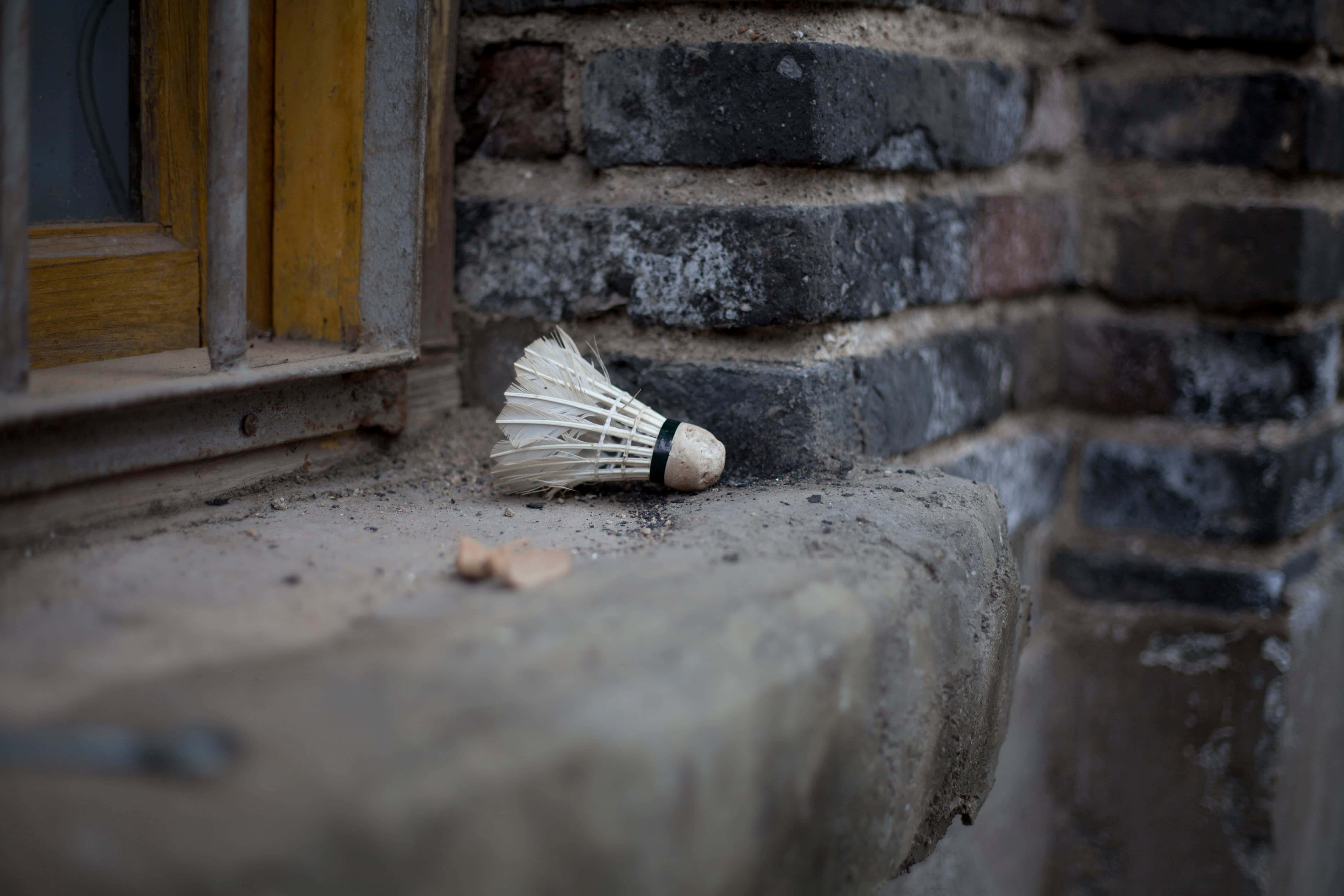 Badminton 桌布 壁紙 — 一個殘舊的羽毛球喺一個風化嘅石窗台上面，旁邊係一堵鄉村風格嘅磚牆同一個黃色框嘅窗。