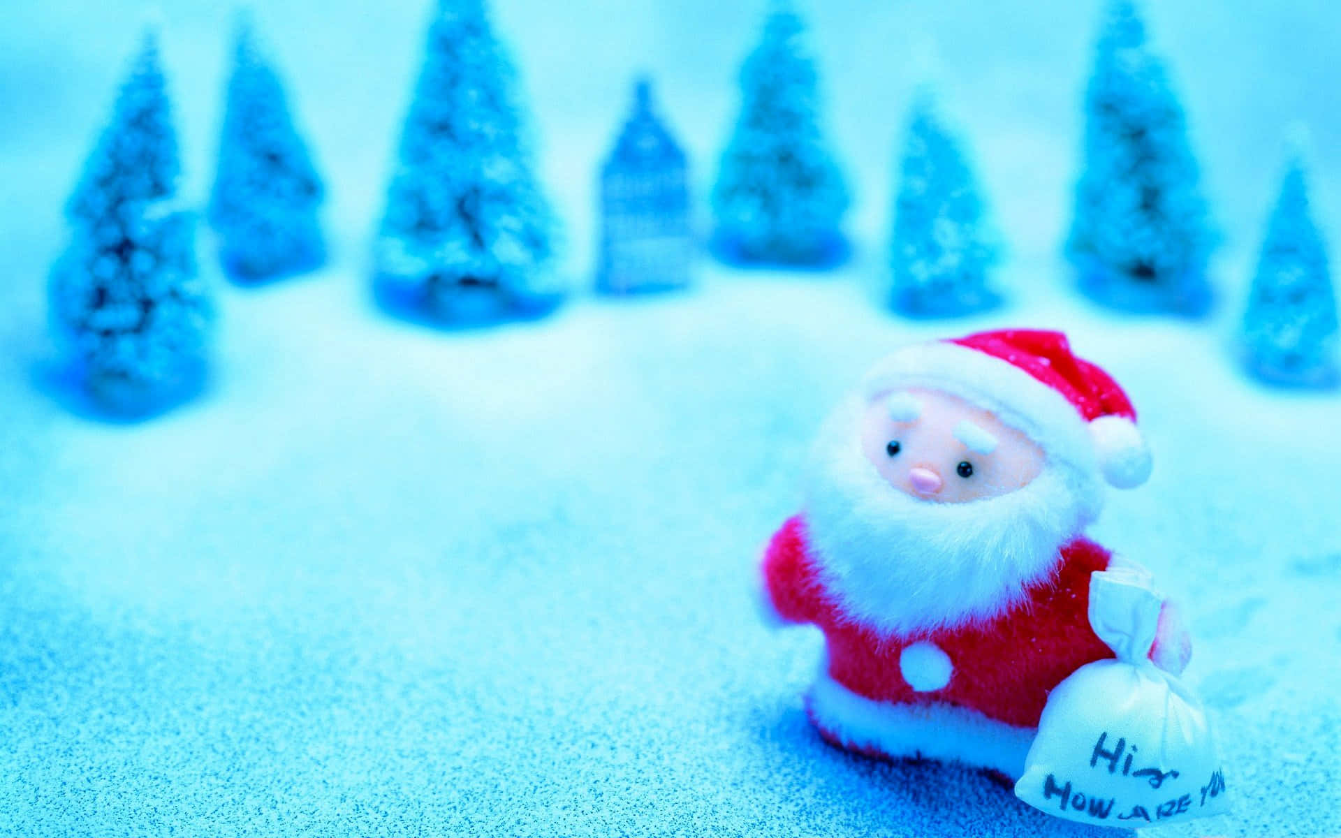 Santa Claus 桌布 壁紙 — 一個毛絨聖誕老人公仔手持手寫禮物袋，站在雪地上，背景有迷你藍色調松樹。