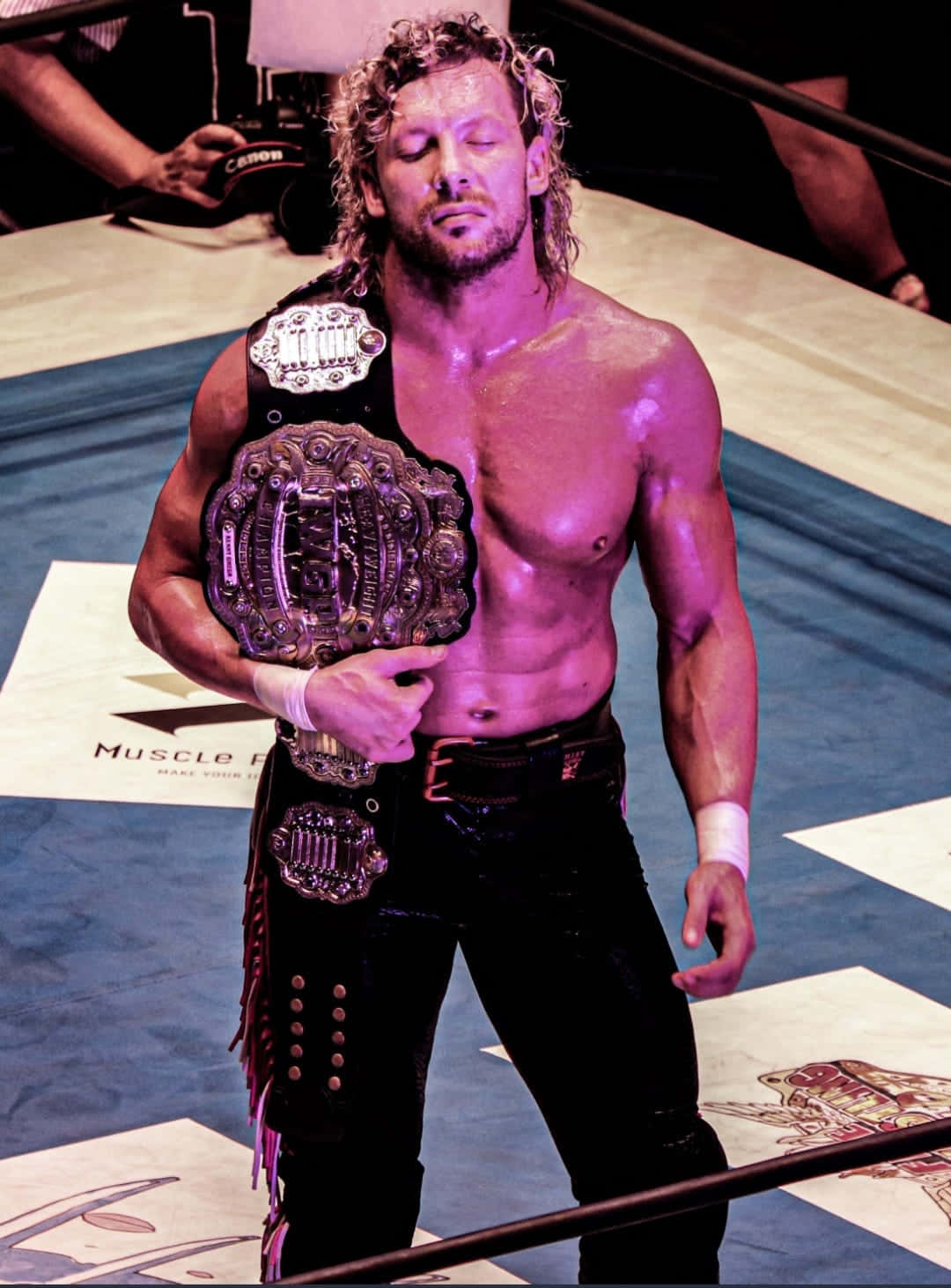 Kenny Omega Eyes Closed Purple Light 壁紙 — 一個汗流浹背、捲髮的摔角手站在擂台上，在紫色舞台燈光下舉著冠軍腰帶。