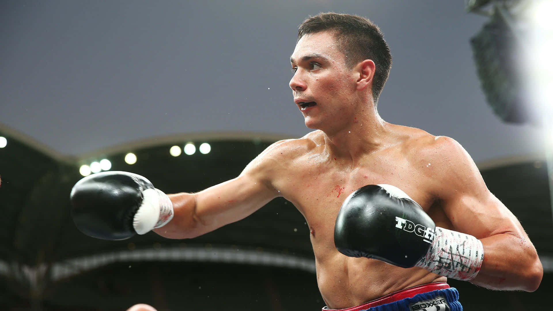 Tim Tszyu 桌布 壁紙 — 一個汗流浹背的男拳擊手戴著黑色手套，在明亮的場館燈光下於一場激烈的比賽中揮拳。