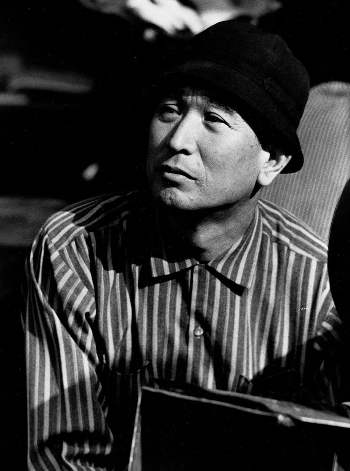 Akira Kurosawa 桌布 壁紙 — 一個沉思中的男人，身穿條紋襯衫和帽子，在光線昏暗、帶有柔和陰影的房間裡若有所思地凝視著。
