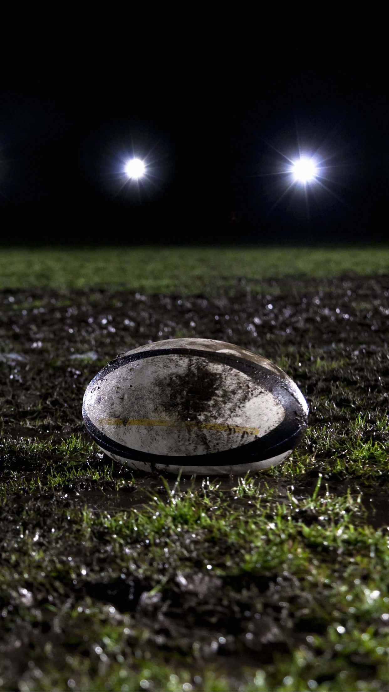 Rugby 桌布 壁紙 — 一個沾滿泥濘的欖球在夜間明亮的球場燈光下放在濕漉漉的草地上。