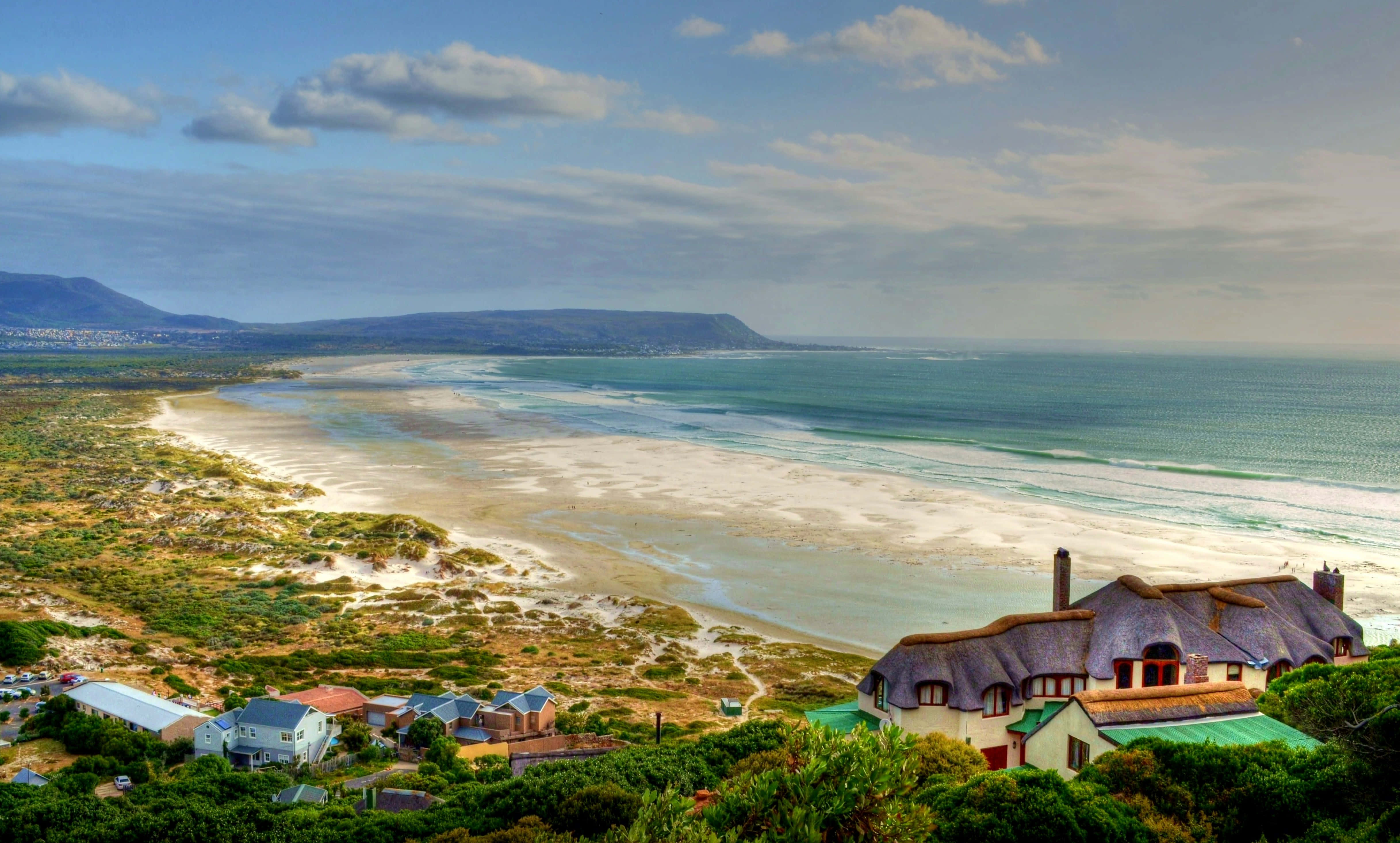 South Africa 桌布 壁紙 — 一個沿海村莊，茅草屋頂的房子坐落在綠色懸崖上，俯瞰著廣闊的沙灘和碧綠的海洋，天空部分多雲。