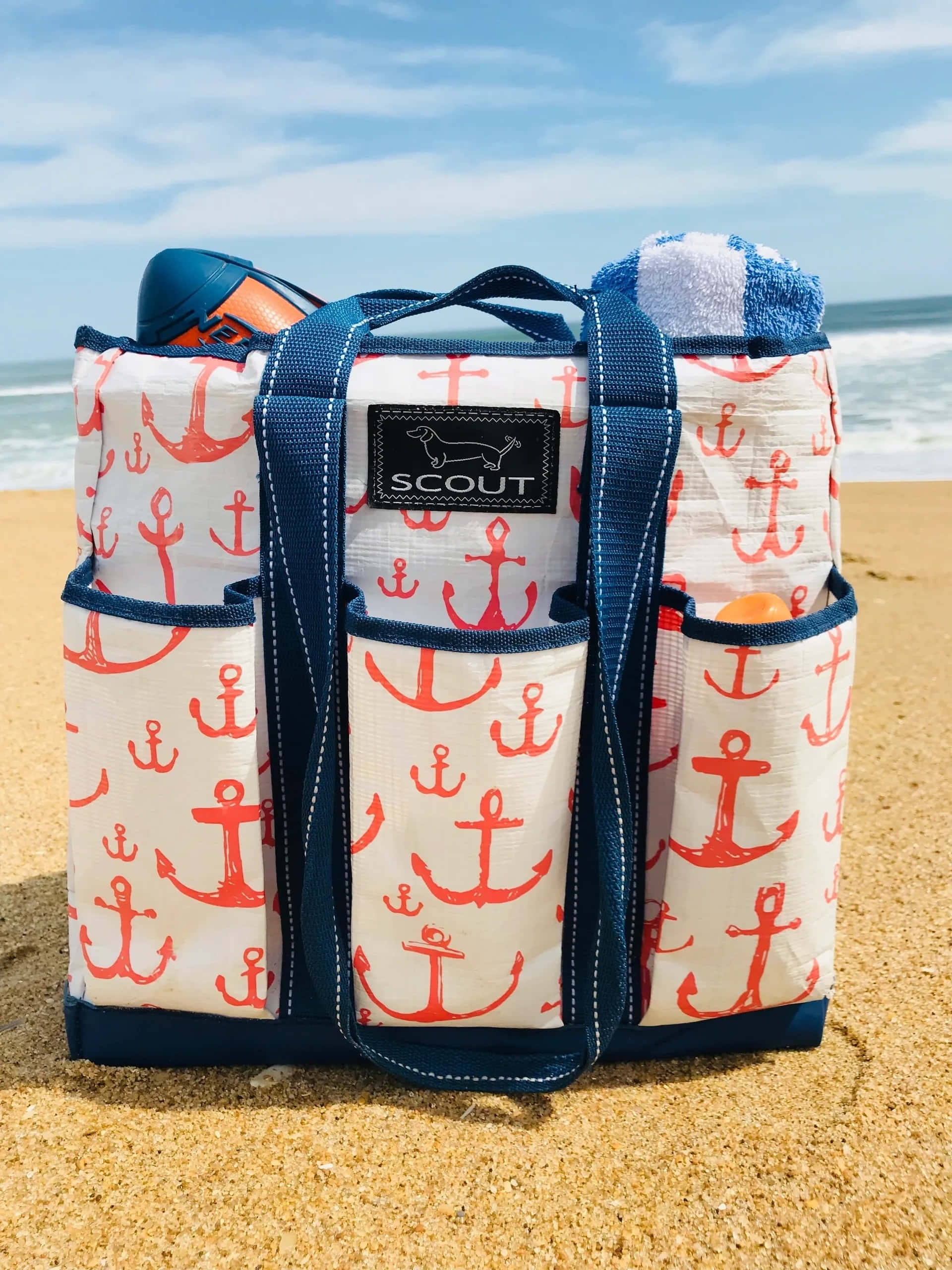 Beach Bag 桌布 壁紙 — 一個海軍藍和珊瑚色船錨圖案沙灘手提袋放在金色沙灘上，背後有海浪和藍天。