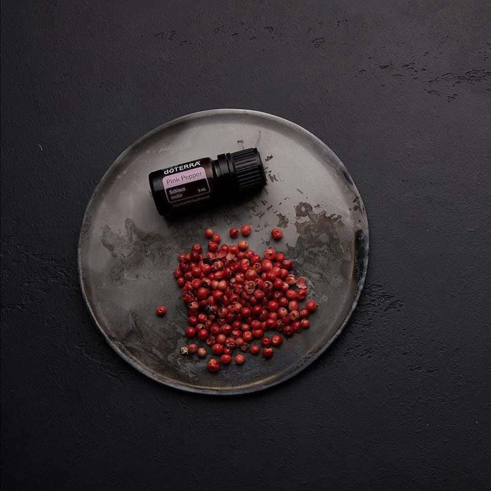 Pink Peppercorn 桌布 壁紙 — 一個深灰色陶瓷盤子上放著鮮豔的紅色粉紅胡椒粒和一個doTERRA精油瓶，背景是帶紋理的黑色。