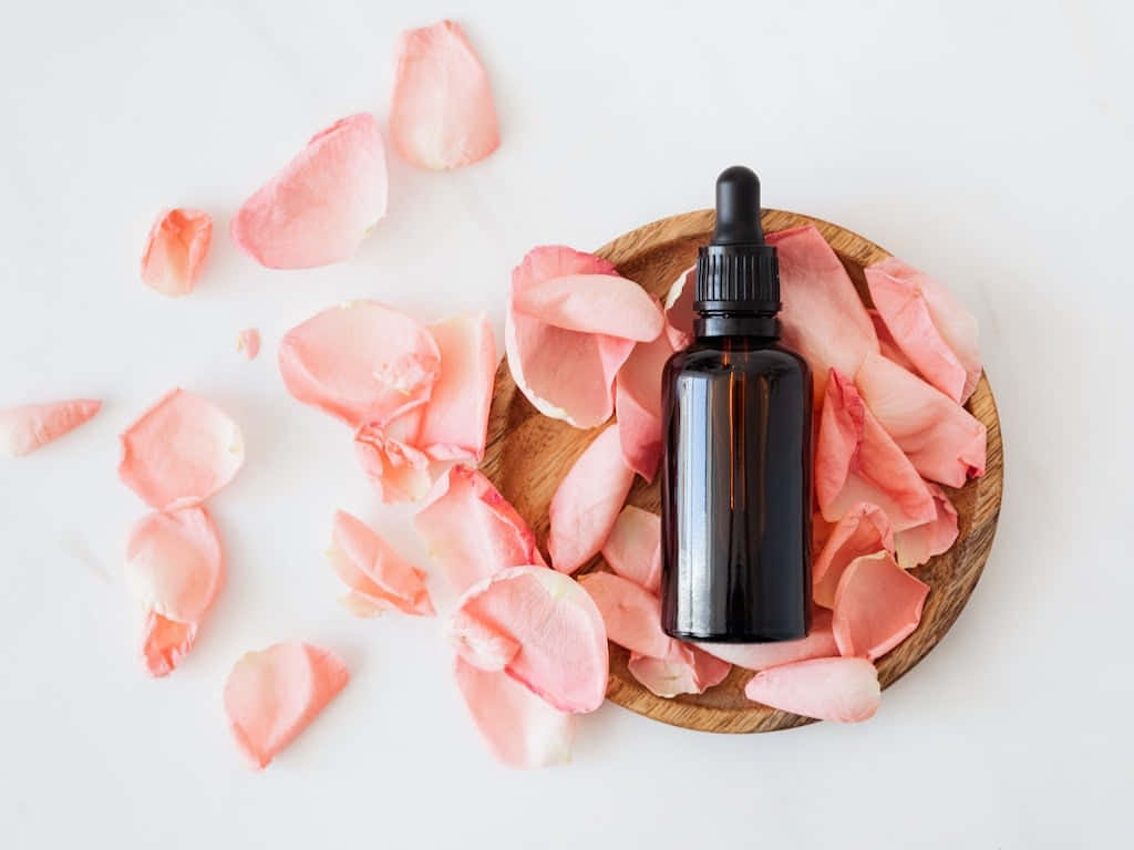 Aromatherapy 桌布 壁紙 — 一個深琥珀色玻璃滴管瓶，放置在木盤中，周圍環繞著柔軟的粉紅玫瑰花瓣，置於潔白表面上。