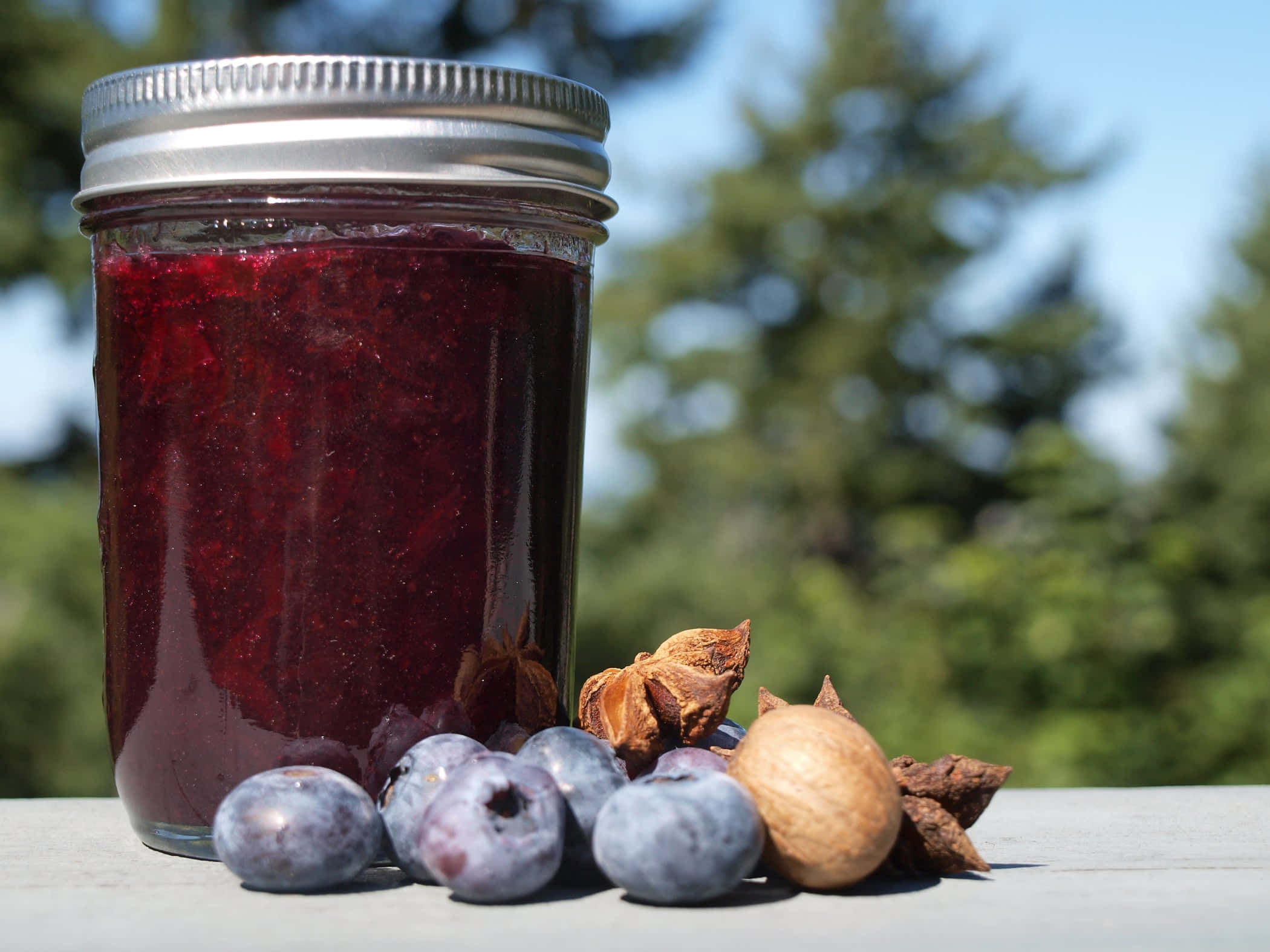Blueberry Jam 桌布 壁紙 — 一個深紅色藍莓醬的玻璃罐放在戶外木桌上，旁邊是新鮮藍莓和八角。