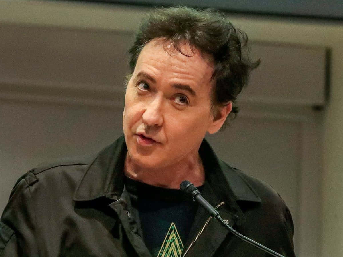 John Cusack 桌布 壁紙 — 一個深色亂髮的男人對著咪高峰說話，身穿黑色外套，內搭帶有綠色幾何圖案的深色襯衫。