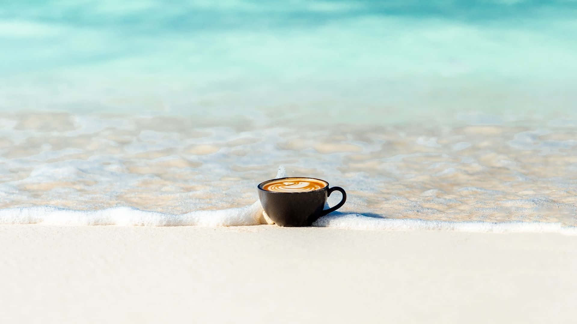 Minimalist Coffee 桌布 壁紙 — 一個深色咖啡杯帶有拉花藝術，放在白色沙灘上，柔和的海浪在明亮的日光下拍打杯底。