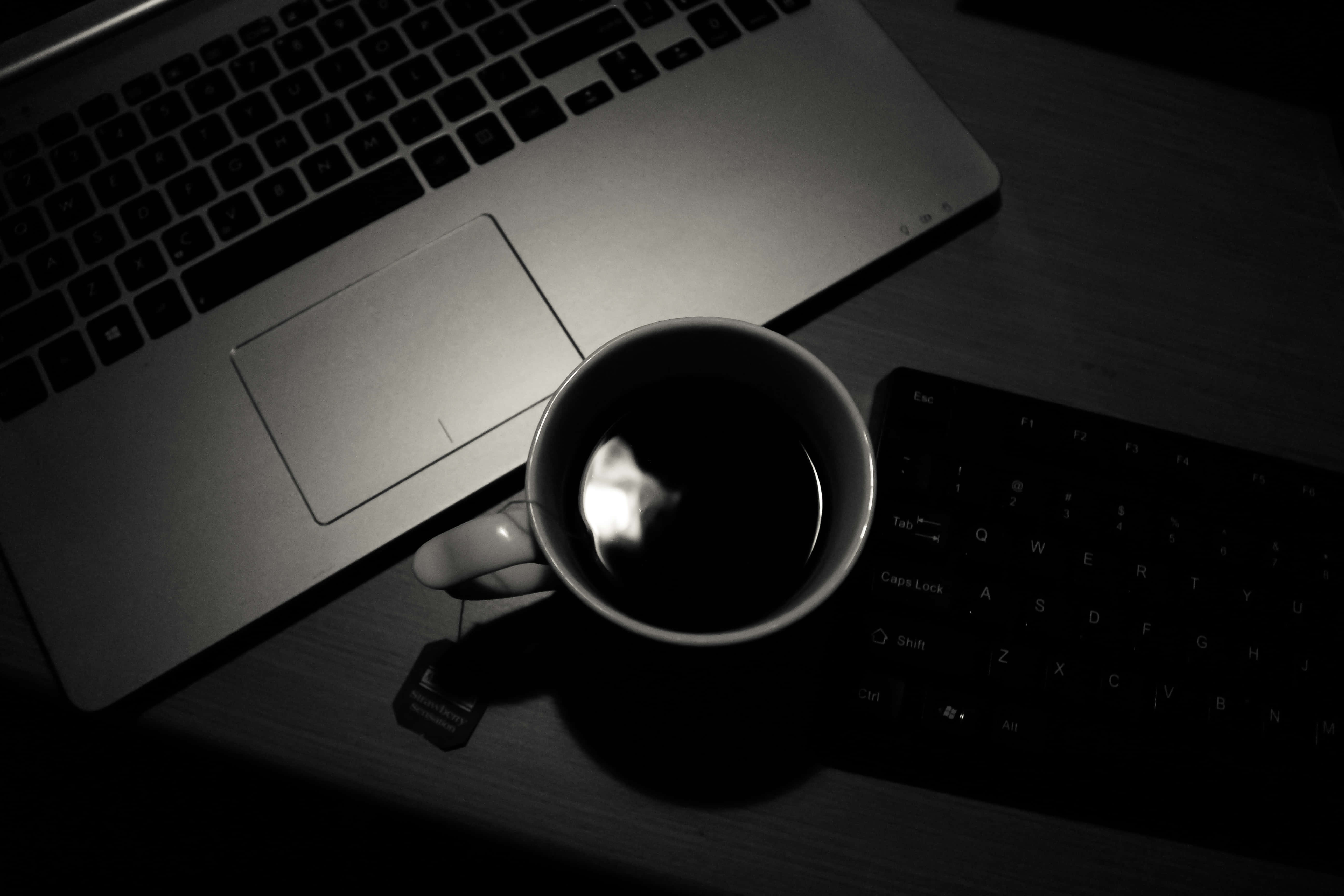 Multimedia 桌布 壁紙 — 一個深色咖啡杯附有茶包，放在筆記型電腦和外接鍵盤旁邊的木桌上，光線昏暗。
