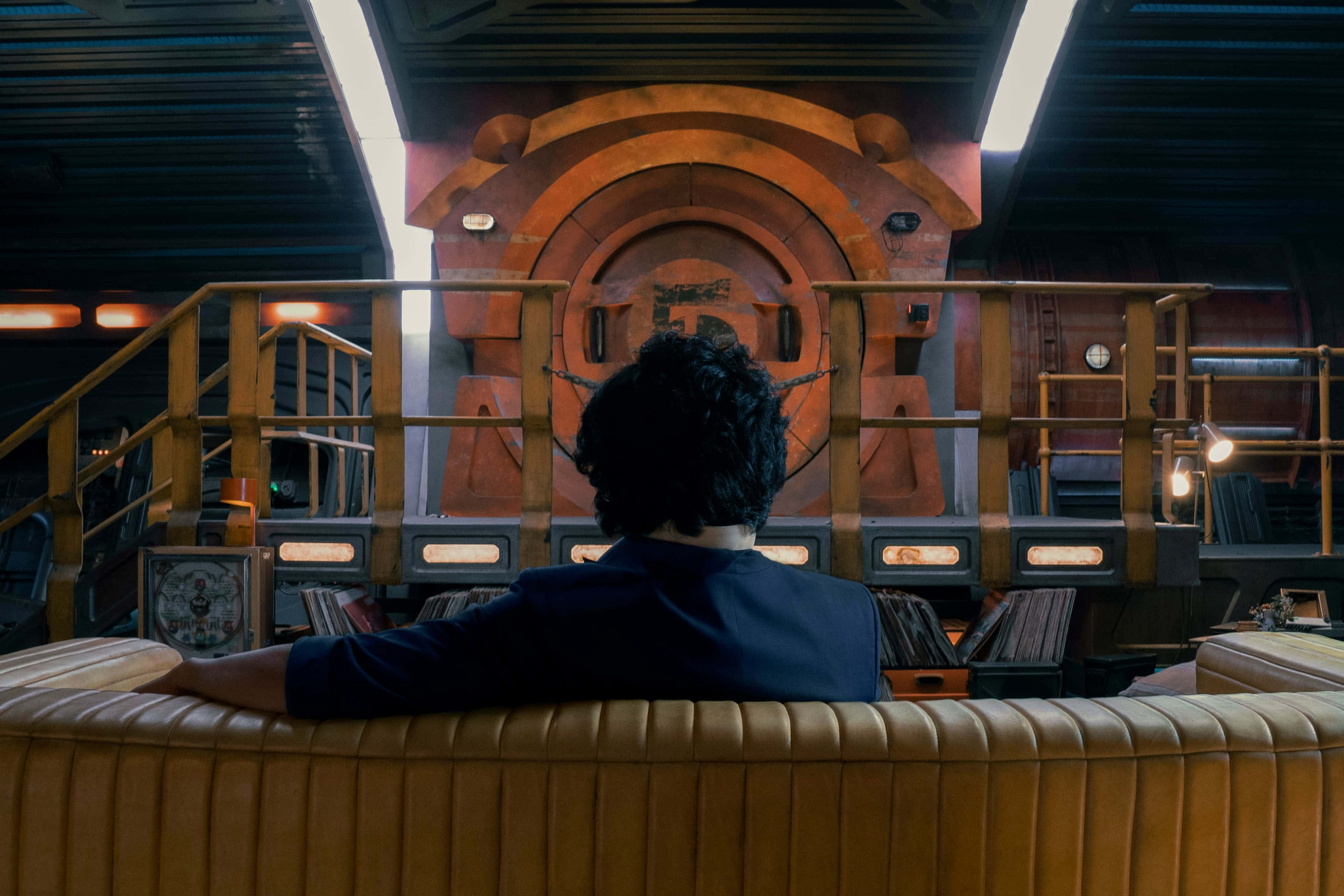 Spike Spiegel 桌布 壁紙 — 一個深色捲髮的人坐在棕色皮梳化上，面向一扇巨大的發光橙色圓形門，身處昏暗的科幻室內。