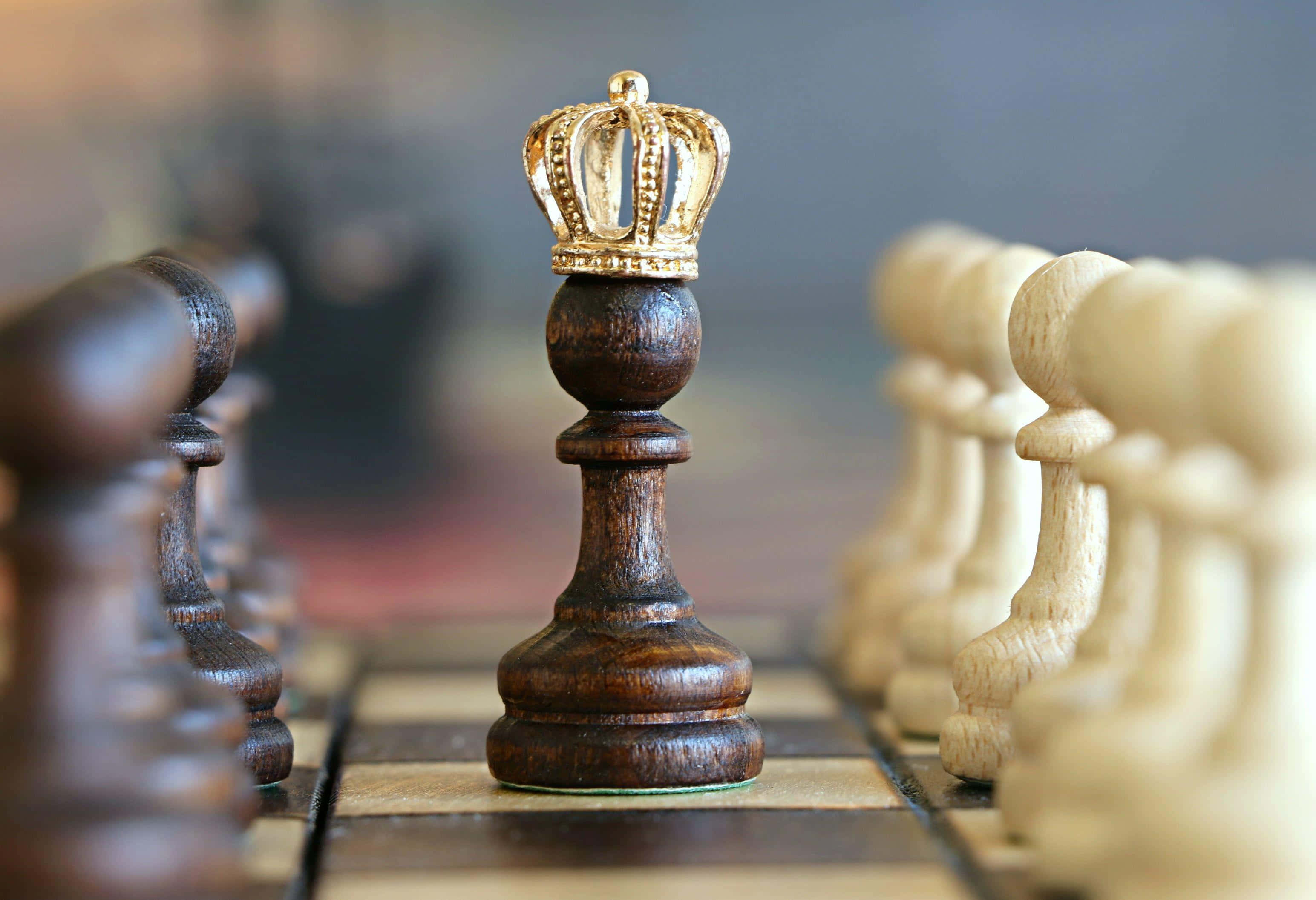 國際象棋 桌布 壁紙 — 一個深色木製國際象棋國王戴上金色王冠的特寫，立於棋盤上，背景有模糊的對手棋子。