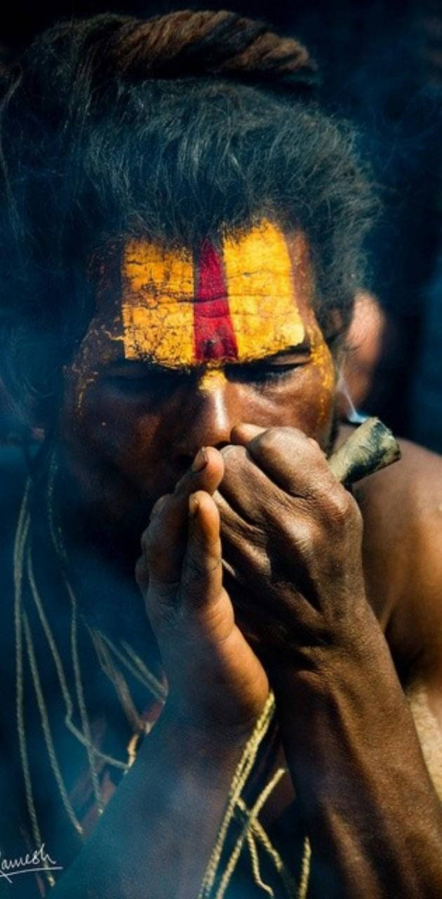Aghori 桌布 壁紙 — 一個深色皮膚、額頭有黃紅色標記的男子在昏暗光線下從煙斗吹出煙霧的特寫。