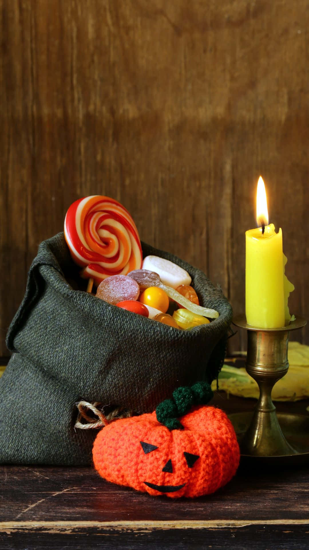 Trick Or Treat Bags 桌布 壁紙 — 一個深色袋子裝滿了色彩繽紛的糖果和一個巨大的漩渦棒棒糖，旁邊是一個鉤針編織的南瓜和一個點燃的黃色蠟燭，放在木桌上。
