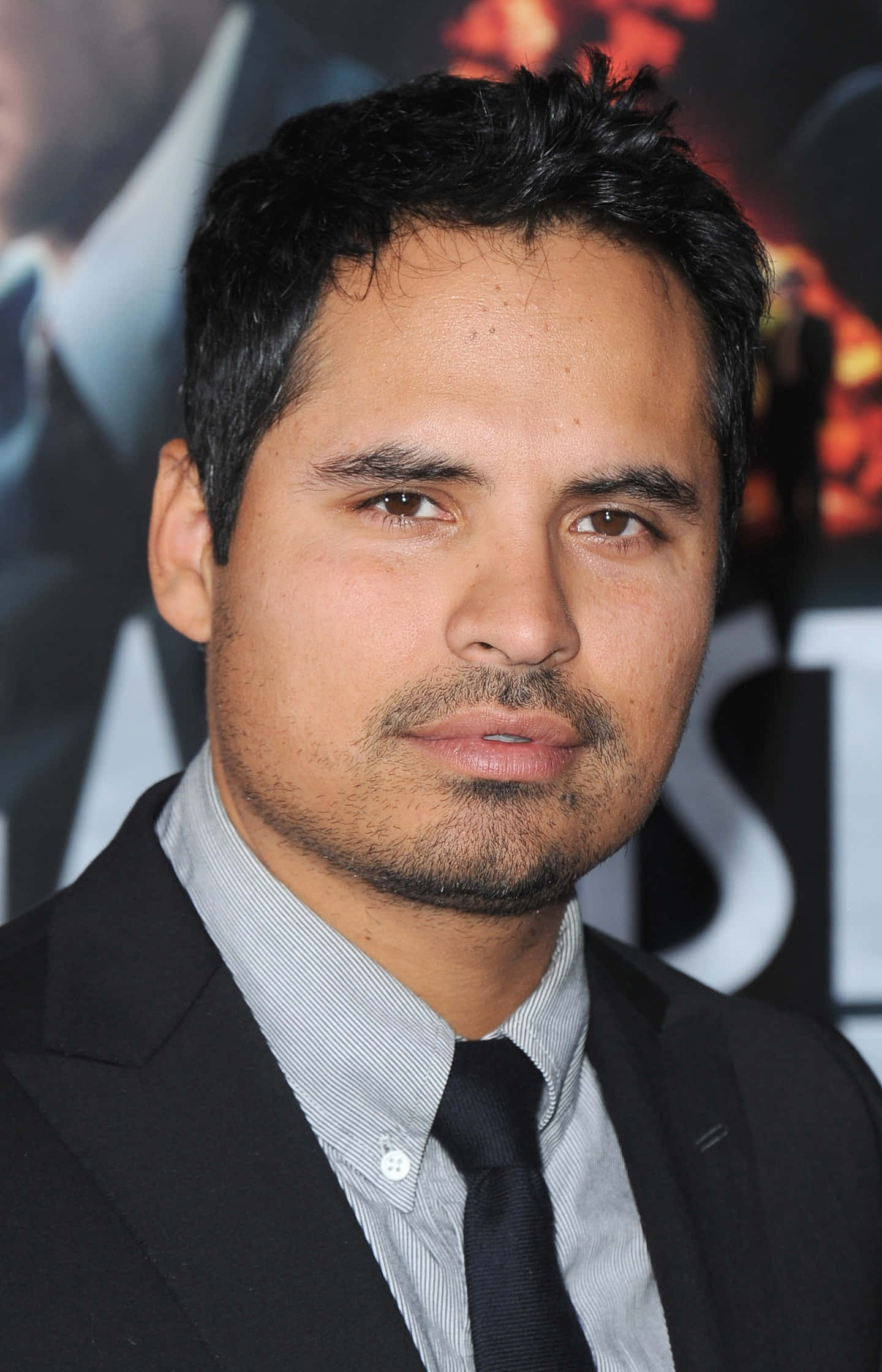Michael Peña 桌布 壁紙 — 一個深色頭髮、留鬚根的男人，身穿黑色西裝打呔，背景是模糊的電影海報，近距離肖像。
