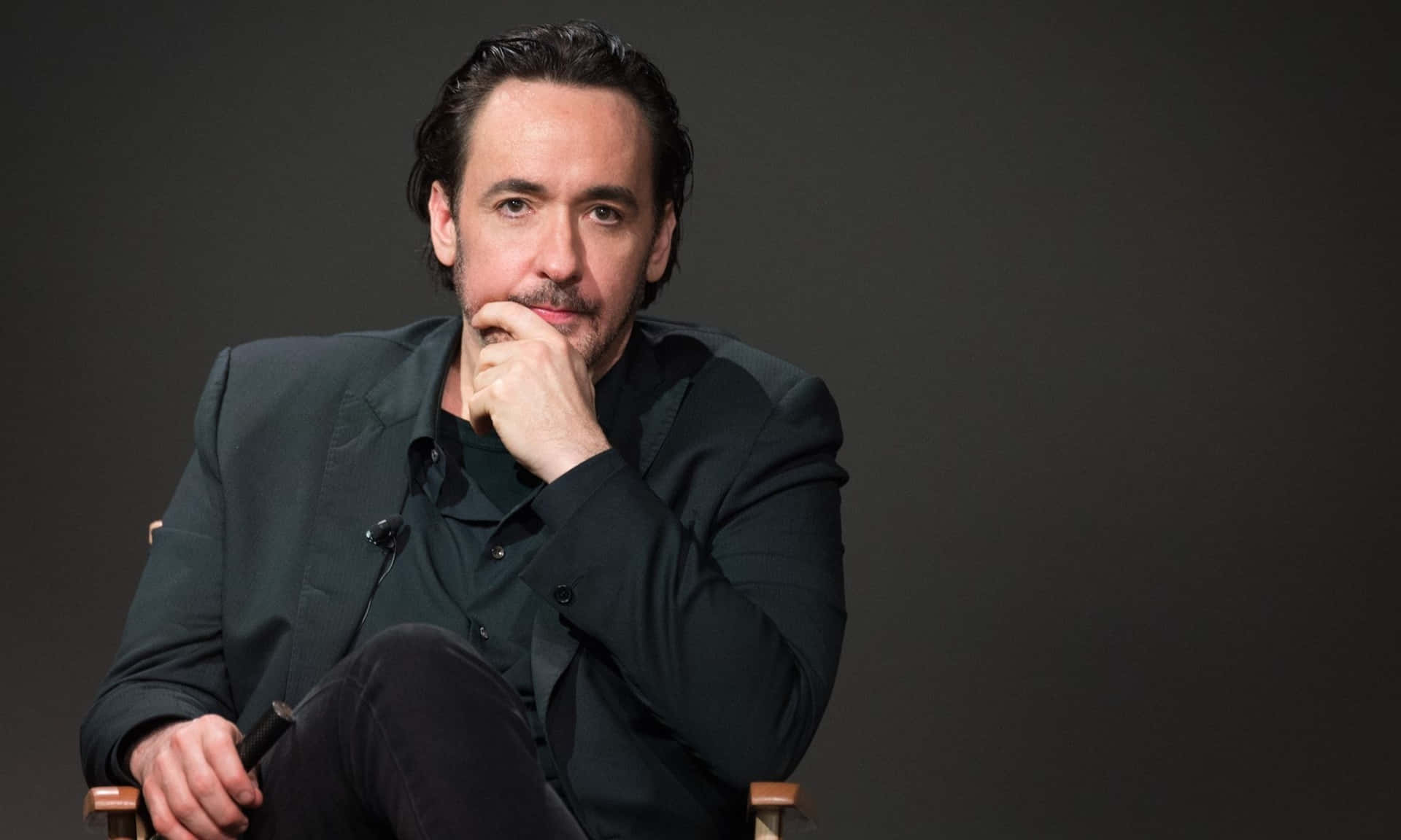 John Cusack 桌布 壁紙 — 一個深色頭髮和鬍鬚的男人坐在導演椅上，若有所思，背景是純深灰色。
