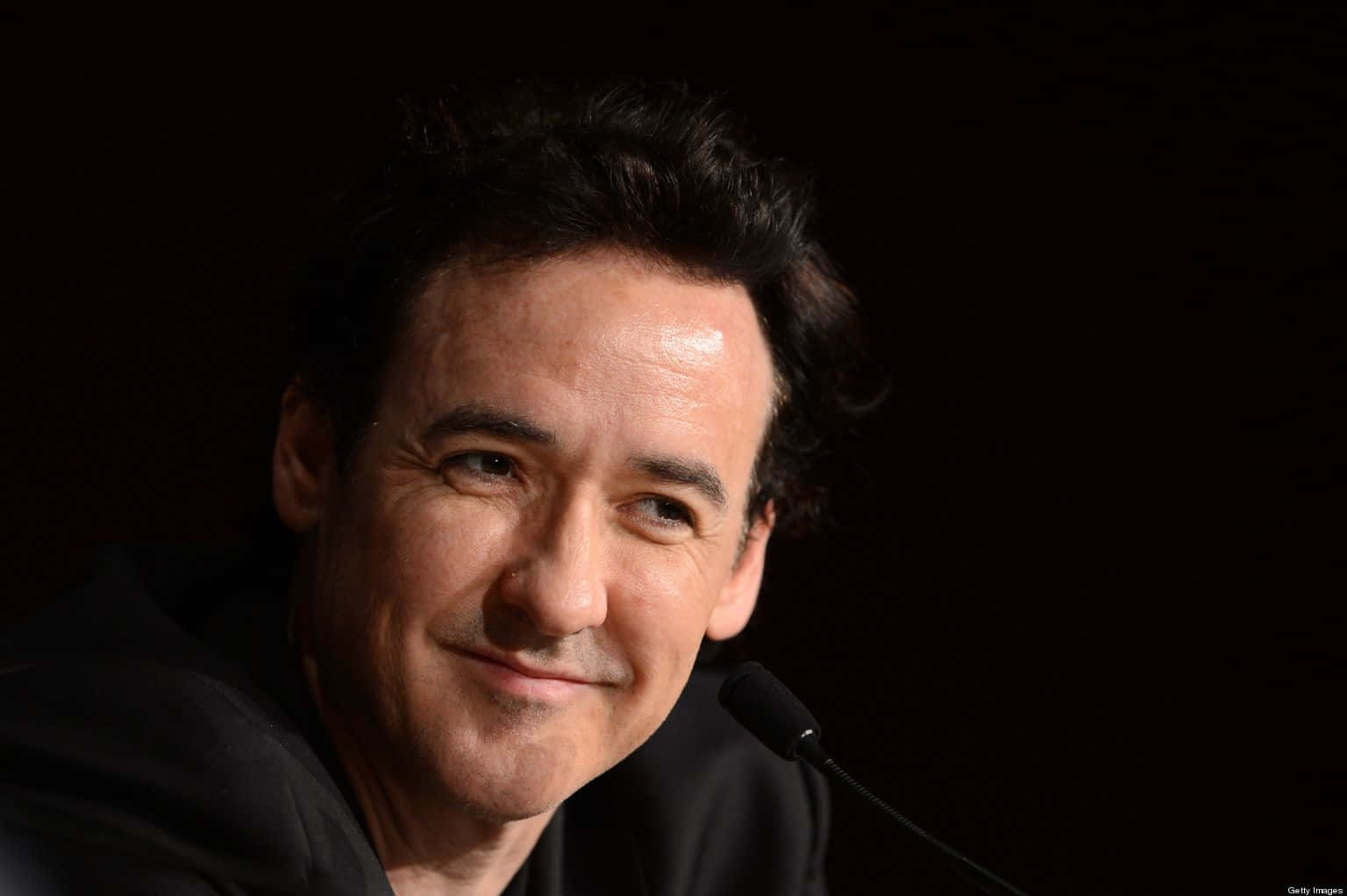 John Cusack 桌布 壁紙 — 一個深色頭髮男士的特寫肖像，在黑色背景前微微微笑，附近有一個麥克風。