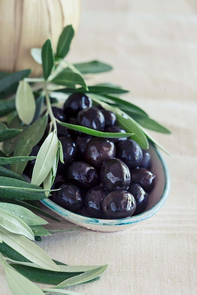 Black Olives 桌布 壁紙 — 一個淺碗盛載著光澤的黑橄欖，放在有紋理的米色布上，旁邊有新鮮的綠色橄欖葉。