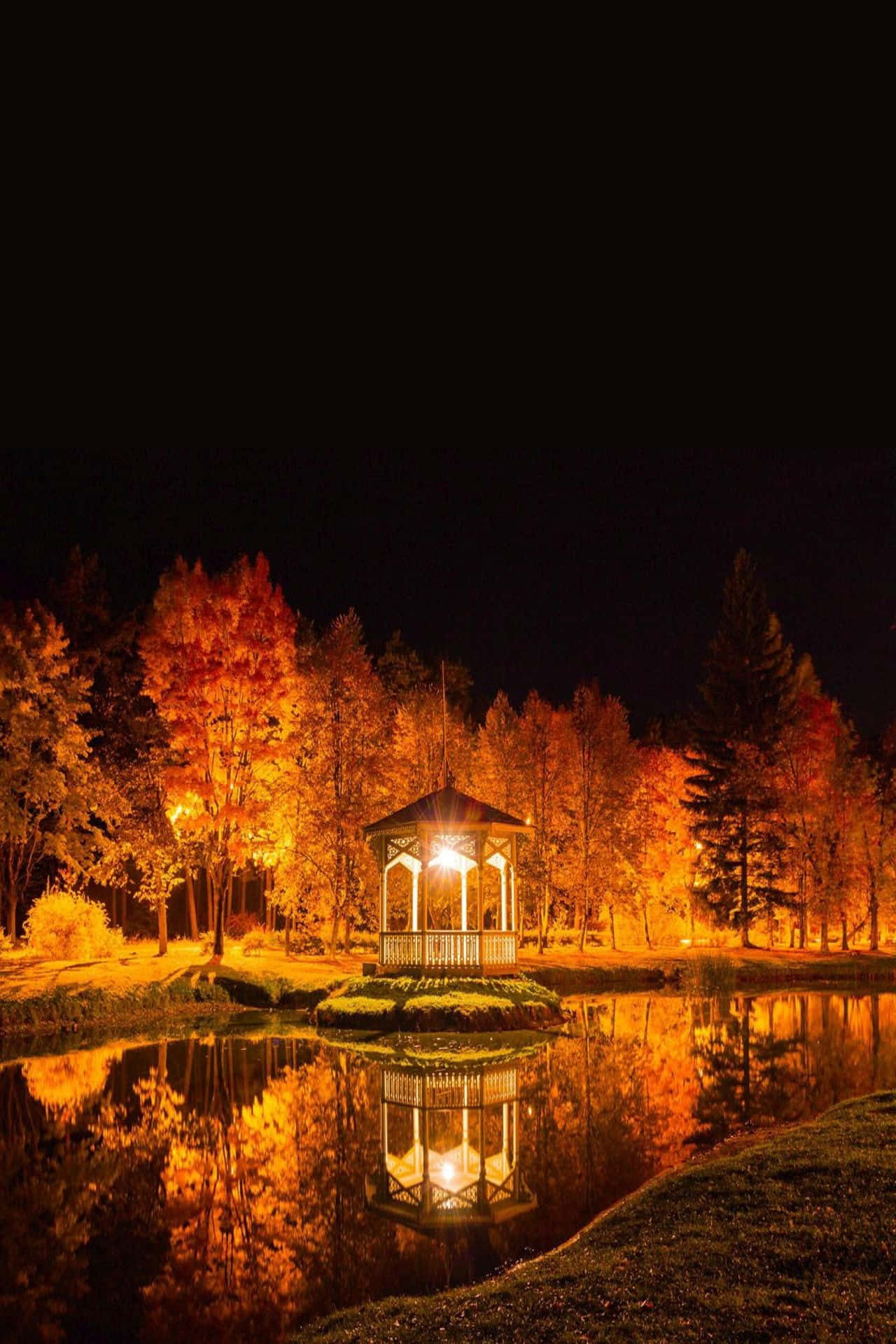 Autumn Evening 桌布 壁紙 — 一個溫暖照明的涼亭在夜間的池塘上發光，其金色倒影映照在平靜的水面上，周圍環繞著秋樹。