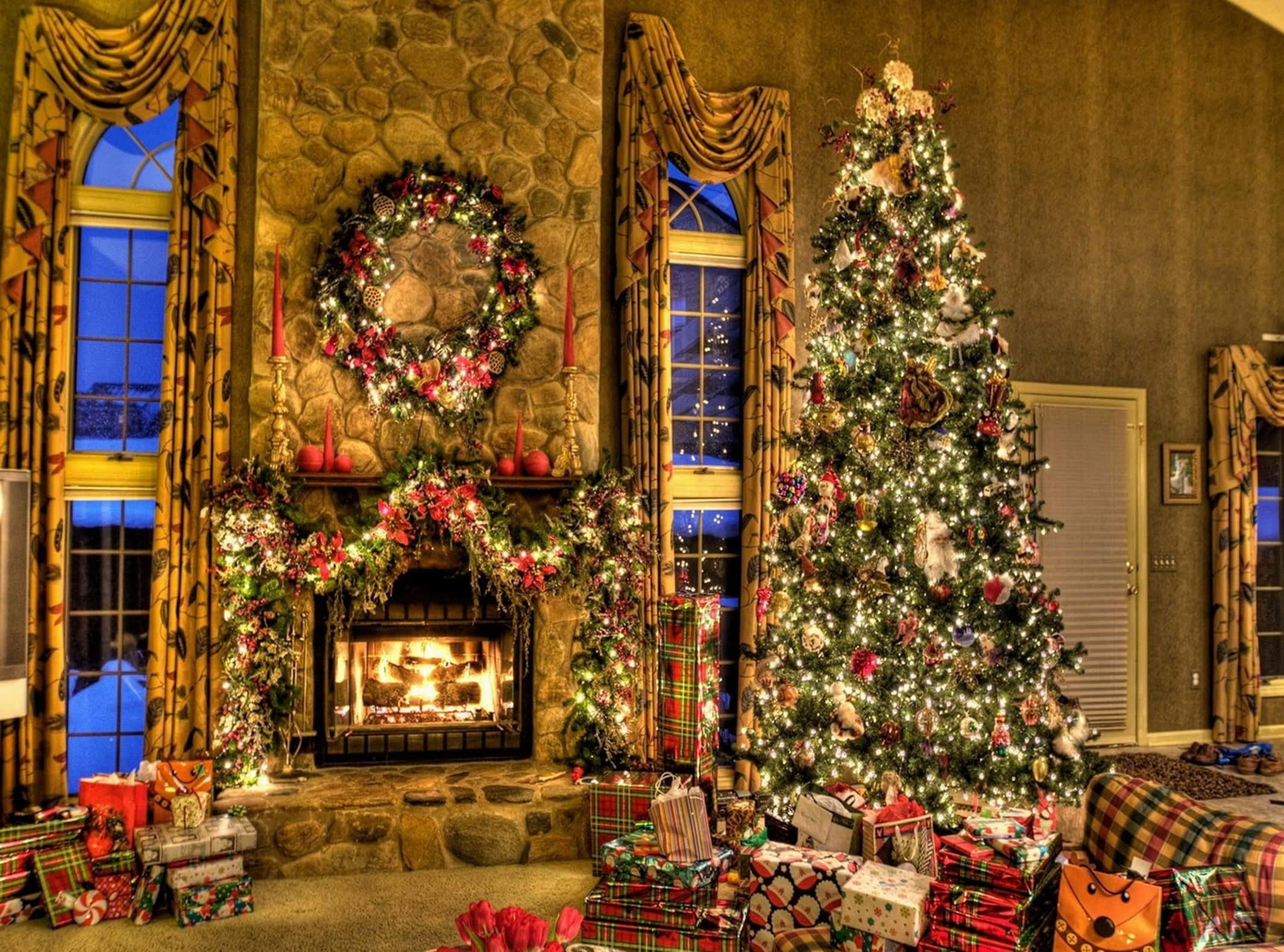 Rustic Christmas 桌布 壁紙 — 一個溫暖照明的石壁爐，裝飾著花卉花環和花彩，旁邊是一棵高聳的聖誕樹，閃爍著燈光和裝飾品，周圍環繞著包裝好的禮物。