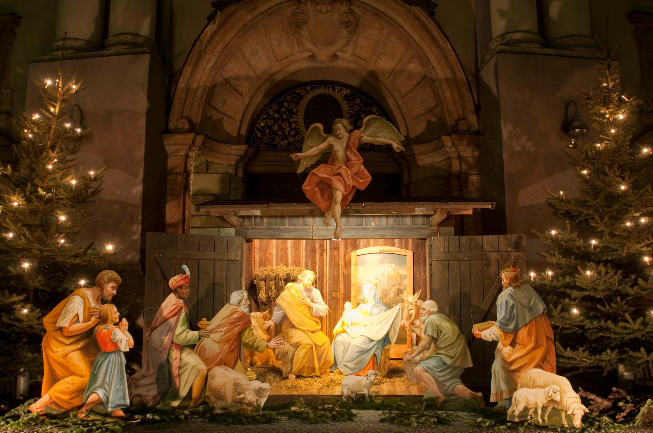 Nativity 桌布 壁紙 — 一個溫暖照明的耶穌誕生場景，包含瑪利亞、約瑟、嬰兒耶穌、牧羊人、東方三博士和天使的雕像，位於拱形石結構下，兩側有裝飾好的聖誕樹。