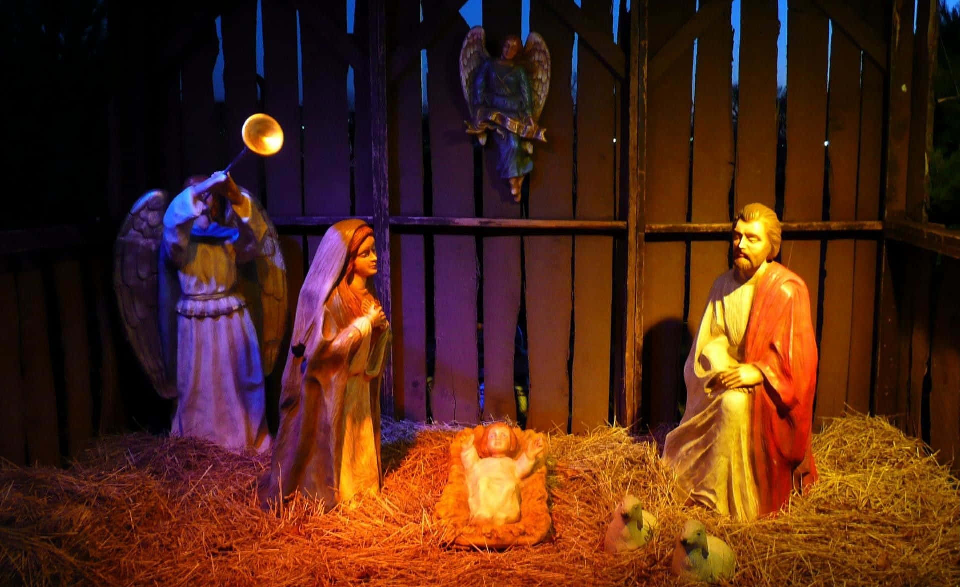 Nativity 桌布 壁紙 — 一個溫暖照明的耶穌誕生場景，有瑪利亞、約瑟、嬰兒耶穌、天使和羊的雕像，在夜間的木製馬廄中。