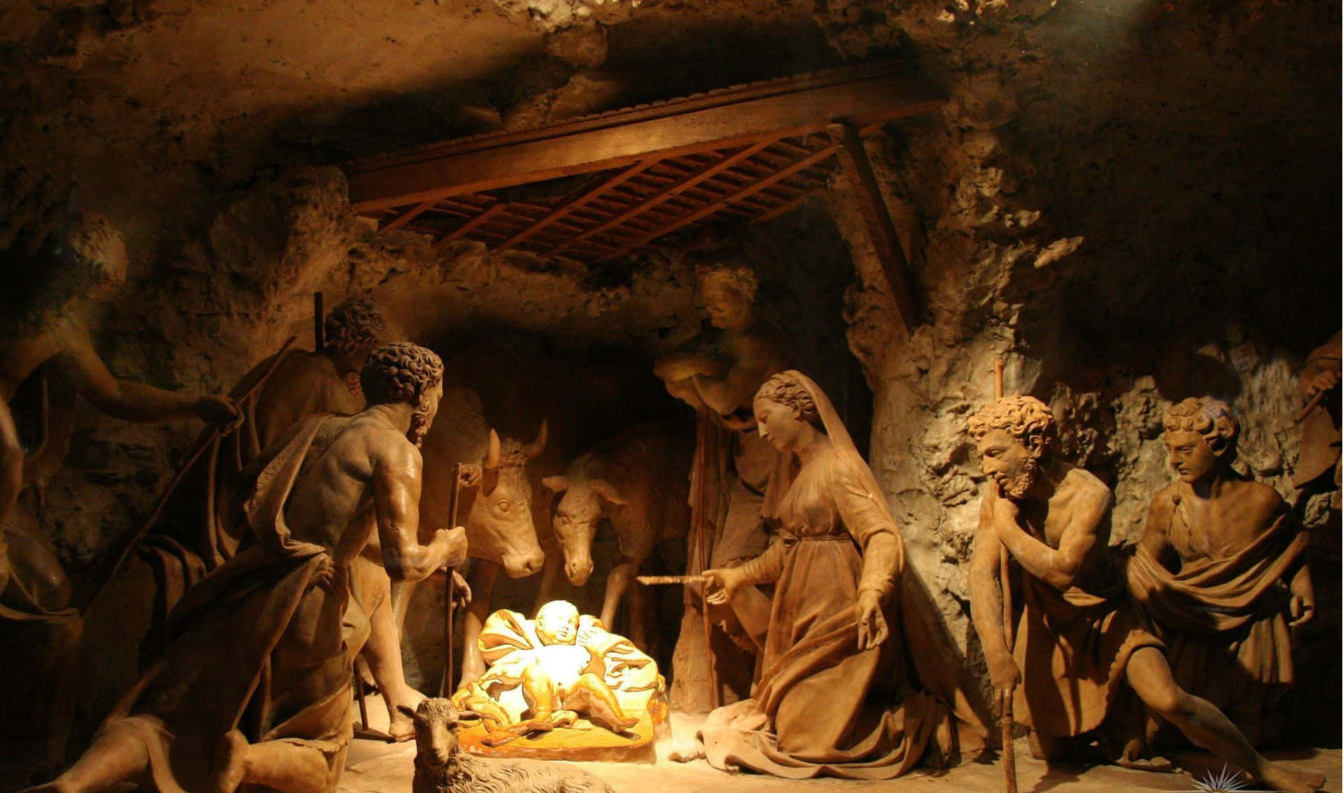 Nativity 桌布 壁紙 — 一個溫暖照明的耶穌誕生場景雕塑，描繪了瑪利亞、約瑟和牧羊人在簡樸的洞穴環境中聚集在嬰兒耶穌周圍。