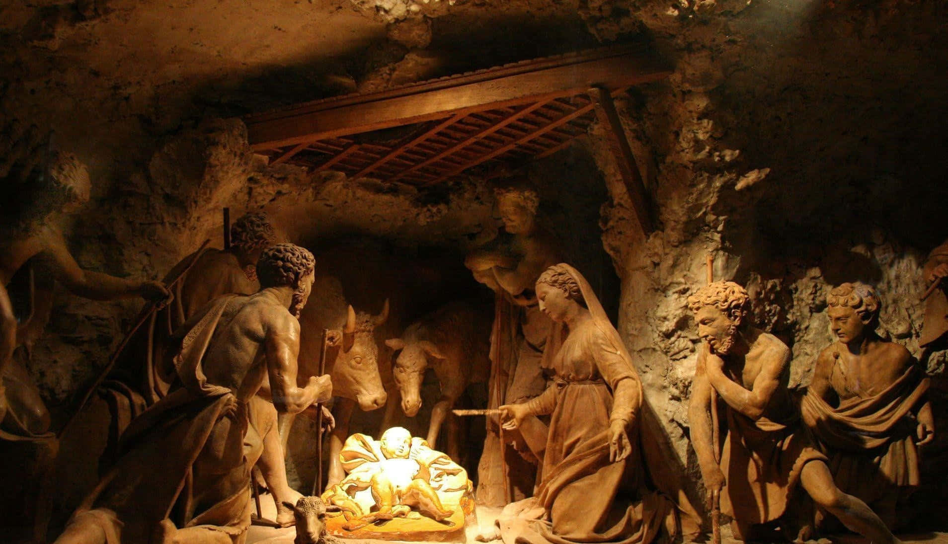 Nativity Scene 桌布 壁紙 — 一個溫暖照明的馬槽場景，瑪利亞、約瑟和牧羊人的石像在質樸的洞穴環境中聚集在嬰兒耶穌周圍。