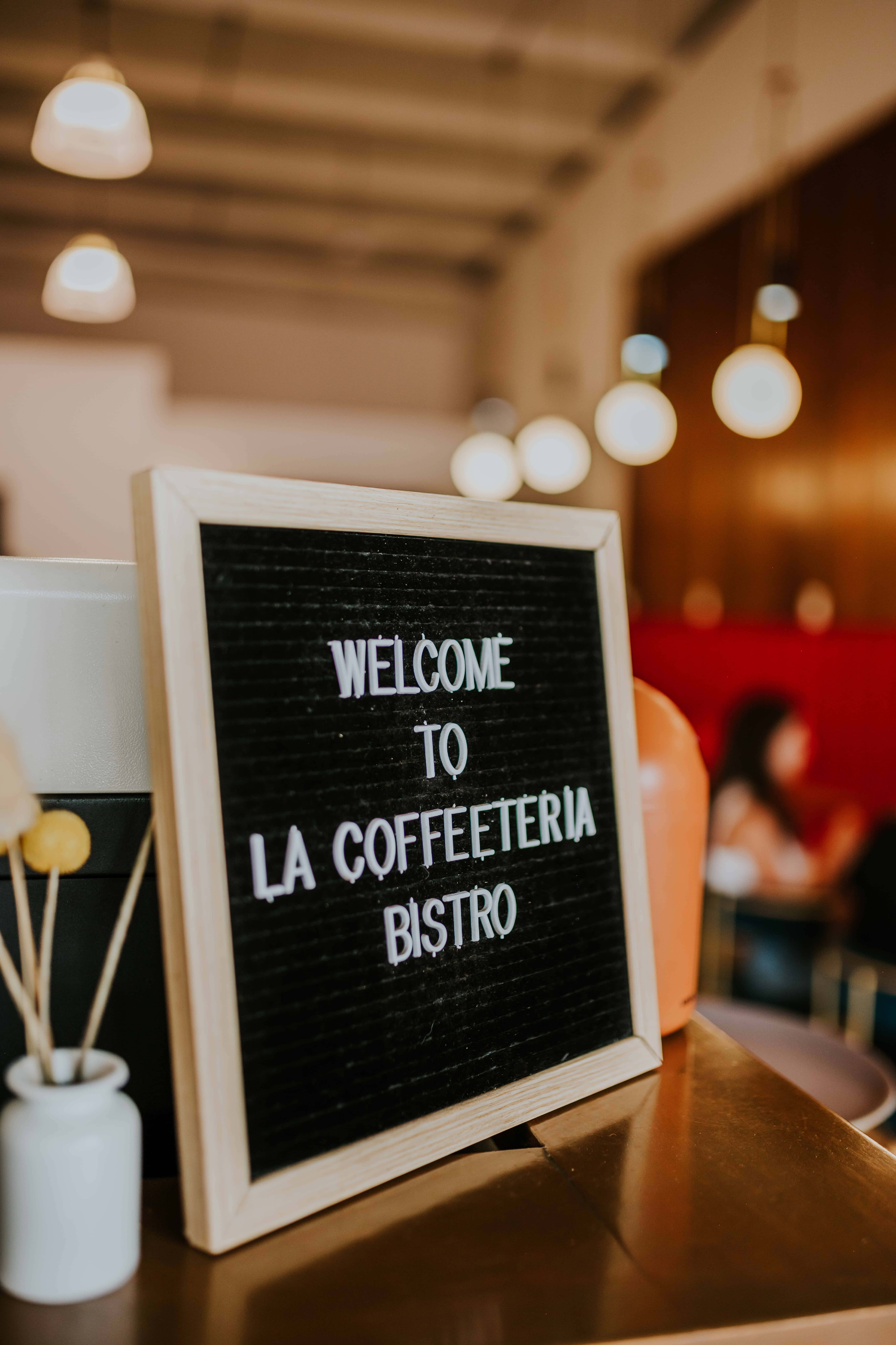 Welcome 桌布 壁紙 — 一個溫暖燈光的咖啡店櫃檯，有一個木框字母板寫著「WELCOME TO LA COFFEETERIA BISTRO」，旁邊有一個白色花瓶插著木棍。
