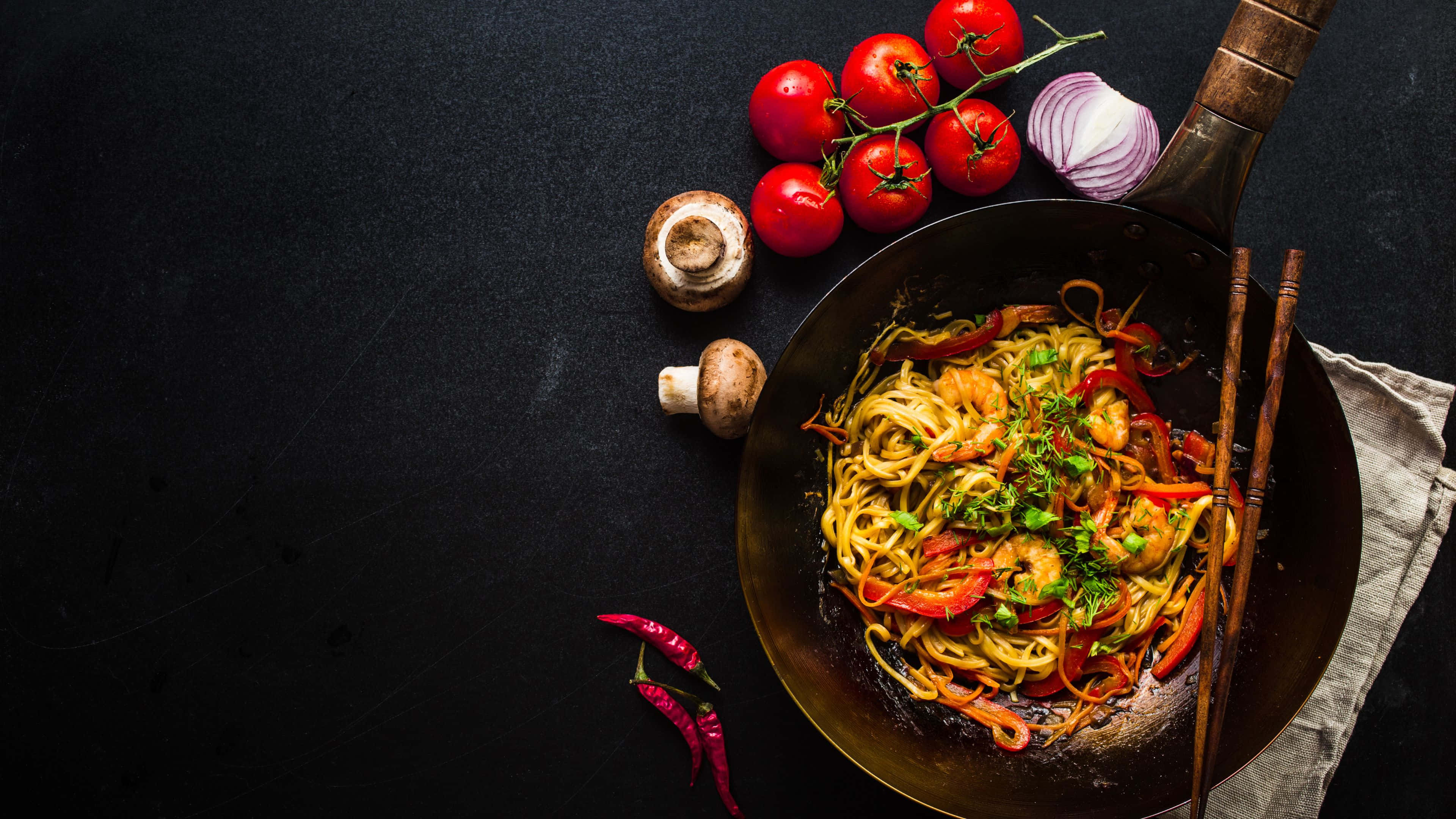 Noodles 桌布 壁紙 — 一個滋滋作響的鑊，裡面有色彩繽紛的炒麵，配上蝦、甜椒和香草，周圍是新鮮番茄、蘑菇和辣椒，放在深色表面上。