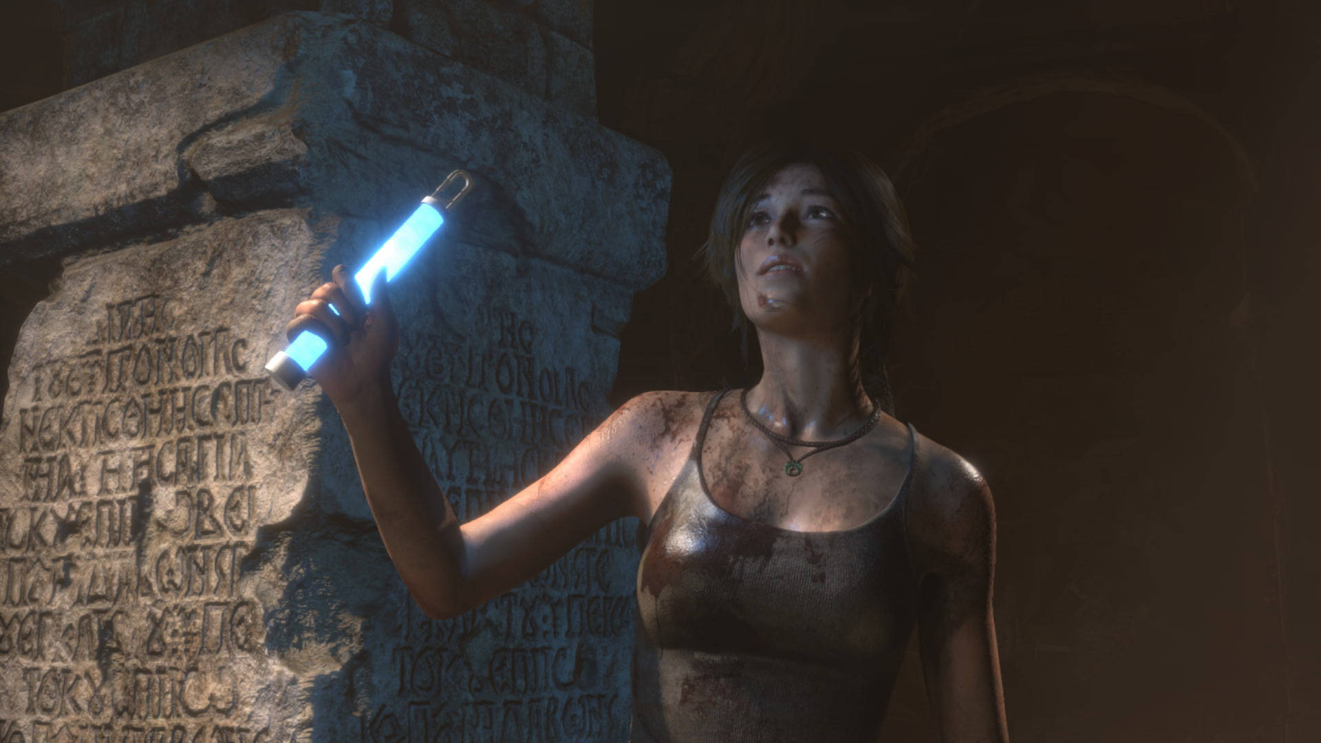 Rise Of The Tomb Raider 桌布 壁紙 — 一個滿身泥濘的冒險家拿著發光的藍色手電筒，照亮黑暗洞穴中的古老雕刻石頭。