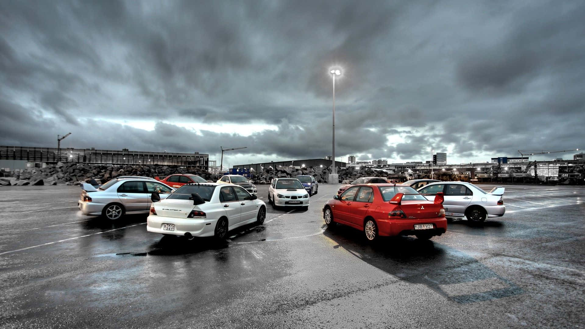 JDM 車 桌布 壁紙 — 一個潮濕的停車場，在暴風雨的天空下，停滿了運動型汽車，包括紅色和白色的三菱Lancer Evos。