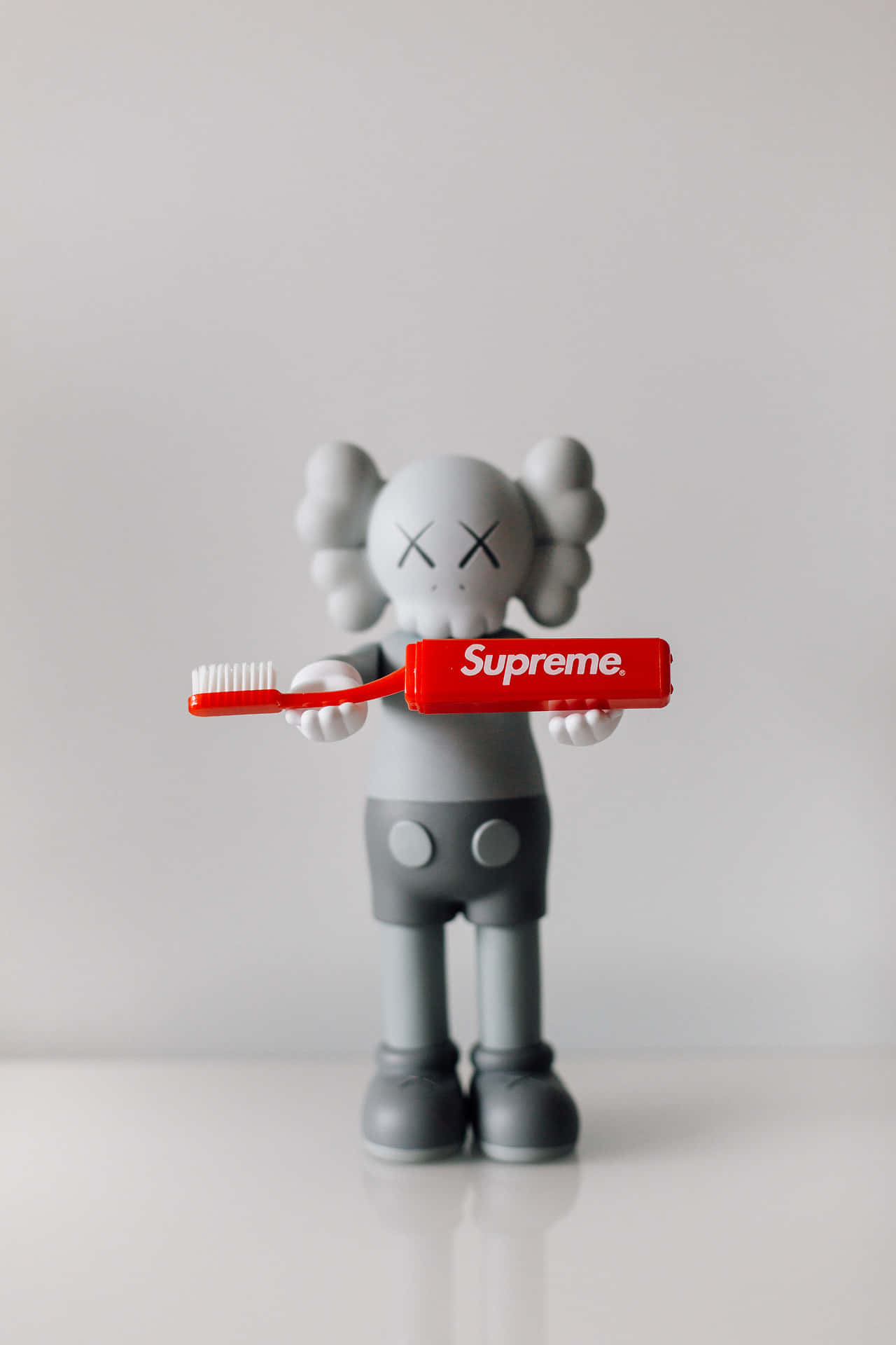Kaws X Supreme 桌布 壁紙 — 一個灰色KAWS公仔喺簡約白色背景前揸住紅色Supreme牙刷。