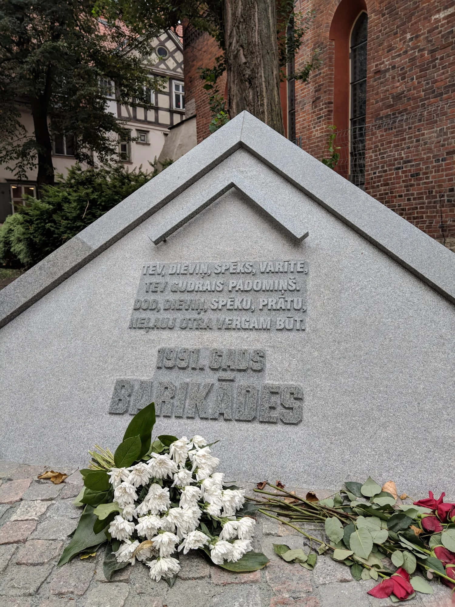 Memorial 桌布 壁紙 — 一個灰色石製紀念碑，上面刻有立陶宛文字和花卉致敬，位於磚砌教堂和樹木旁的鵝卵石小徑上。