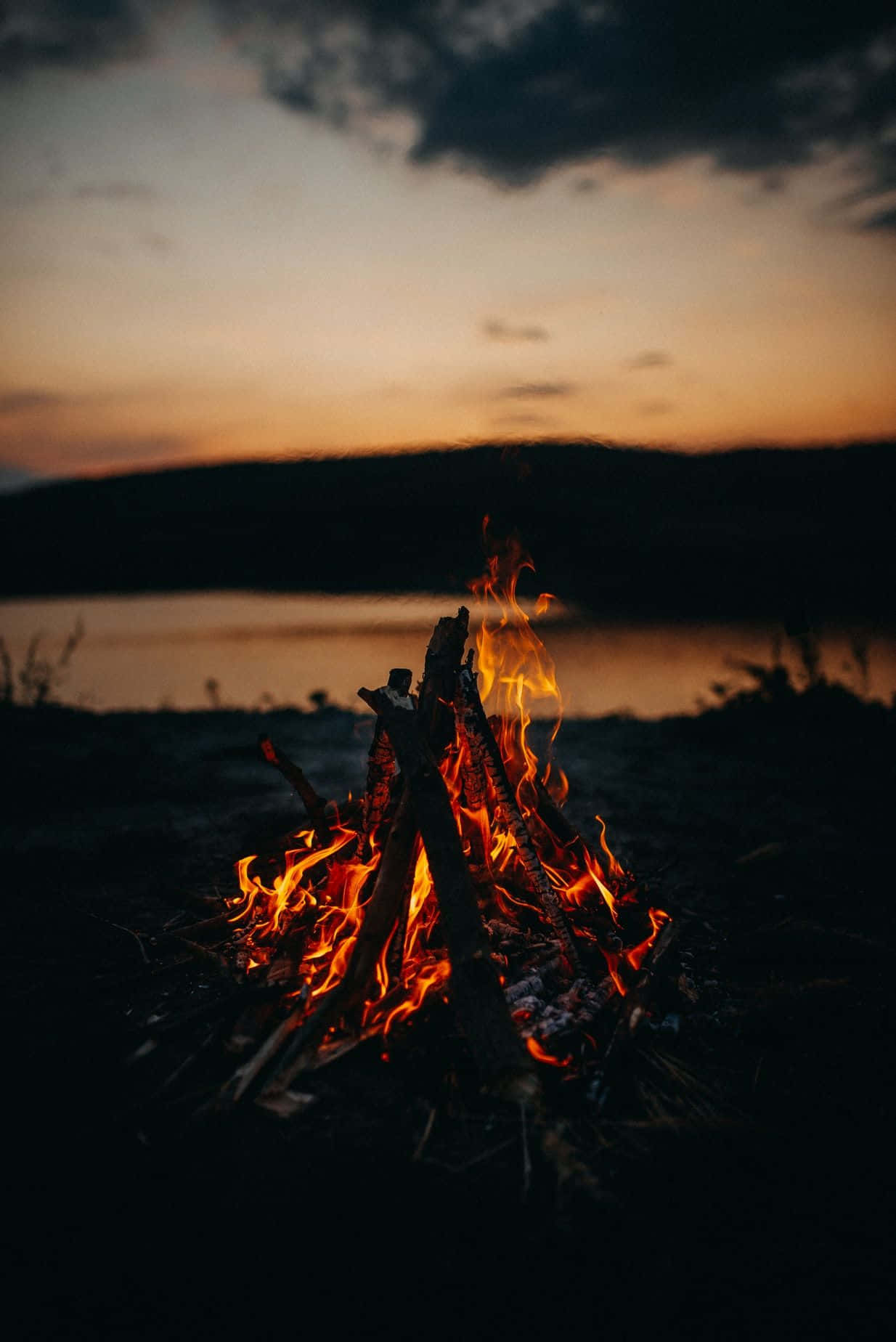 Fall Bonfire 桌布 壁紙 — 一個熊熊燃燒的營火在平靜的湖面上方、遠山環繞的暮色天空中溫暖地發光。