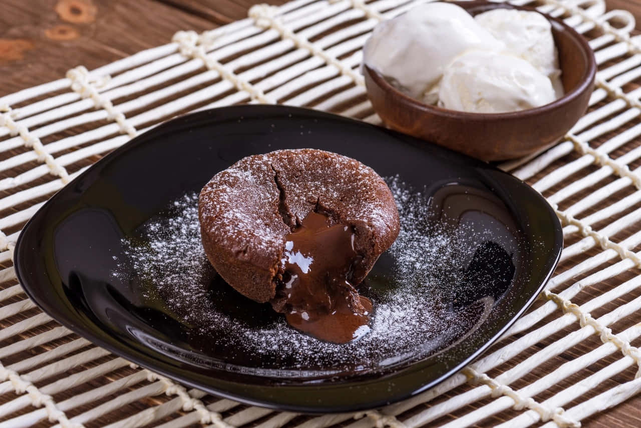 Winter Desserts 桌布 壁紙 — 一個熔岩朱古力蛋糕流出濃郁黏稠的朱古力醬到一個灑滿糖粉的黑色碟子上，旁邊放著一碗雲呢拿雪糕。