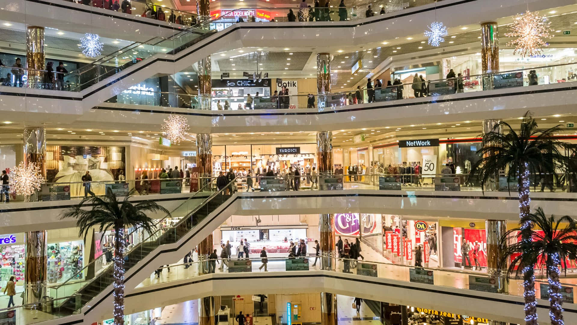 Mall 桌布 壁紙 — 一個熙熙攘攘的多層購物中心，有發光的水晶吊燈、棕櫚樹和顧客在玻璃圍成的自動扶梯之間穿梭。