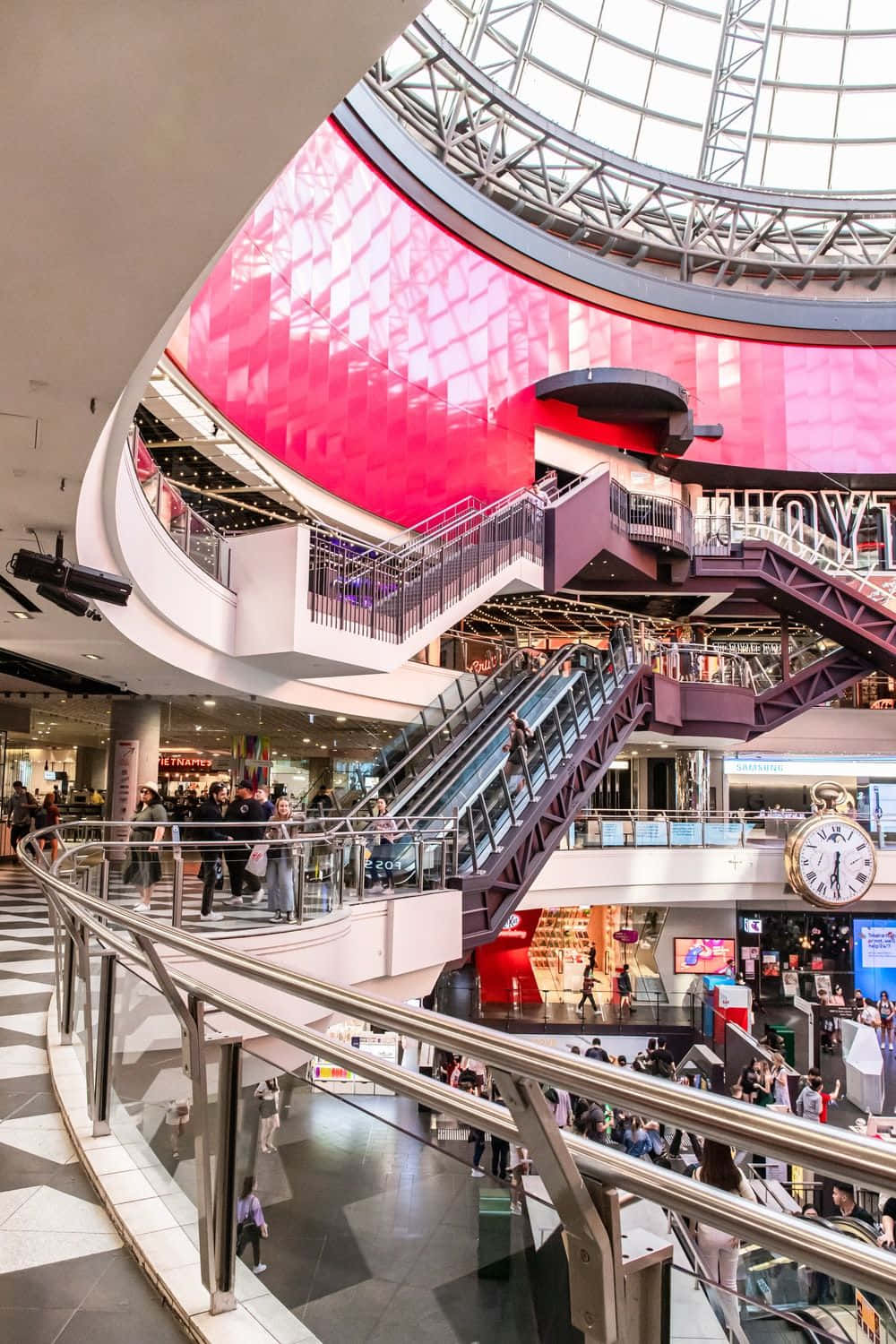 Shopping 桌布 壁紙 — 一個熙熙攘攘的多層購物中心，設有弧形自動扶梯和玻璃穹頂下充滿活力的粉紅色弧形牆。
