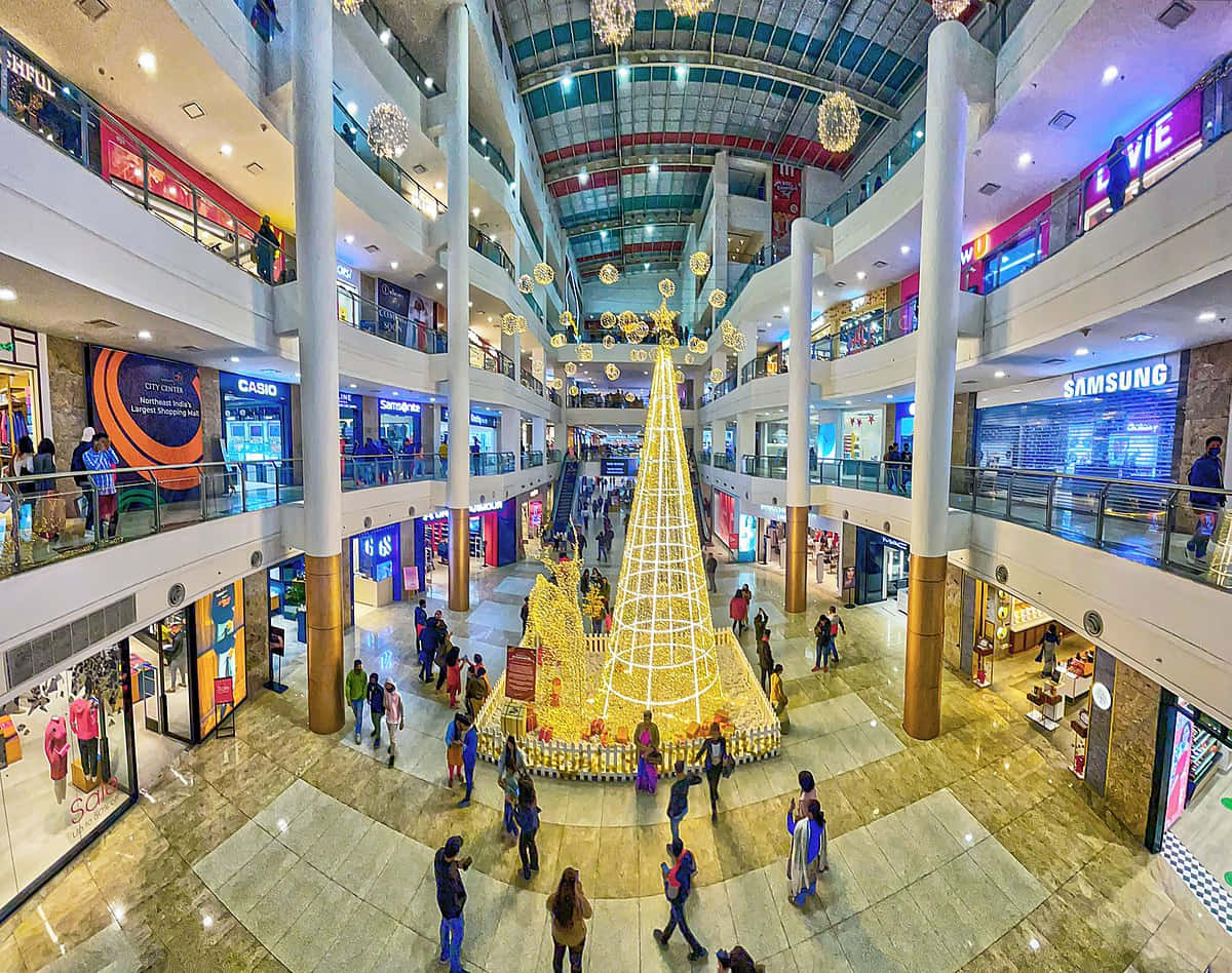 Mall 桌布 壁紙 — 一個熙熙攘攘的多層購物中心中庭，以一座裝飾著燈飾和飾品的金色聖誕樹為中心。