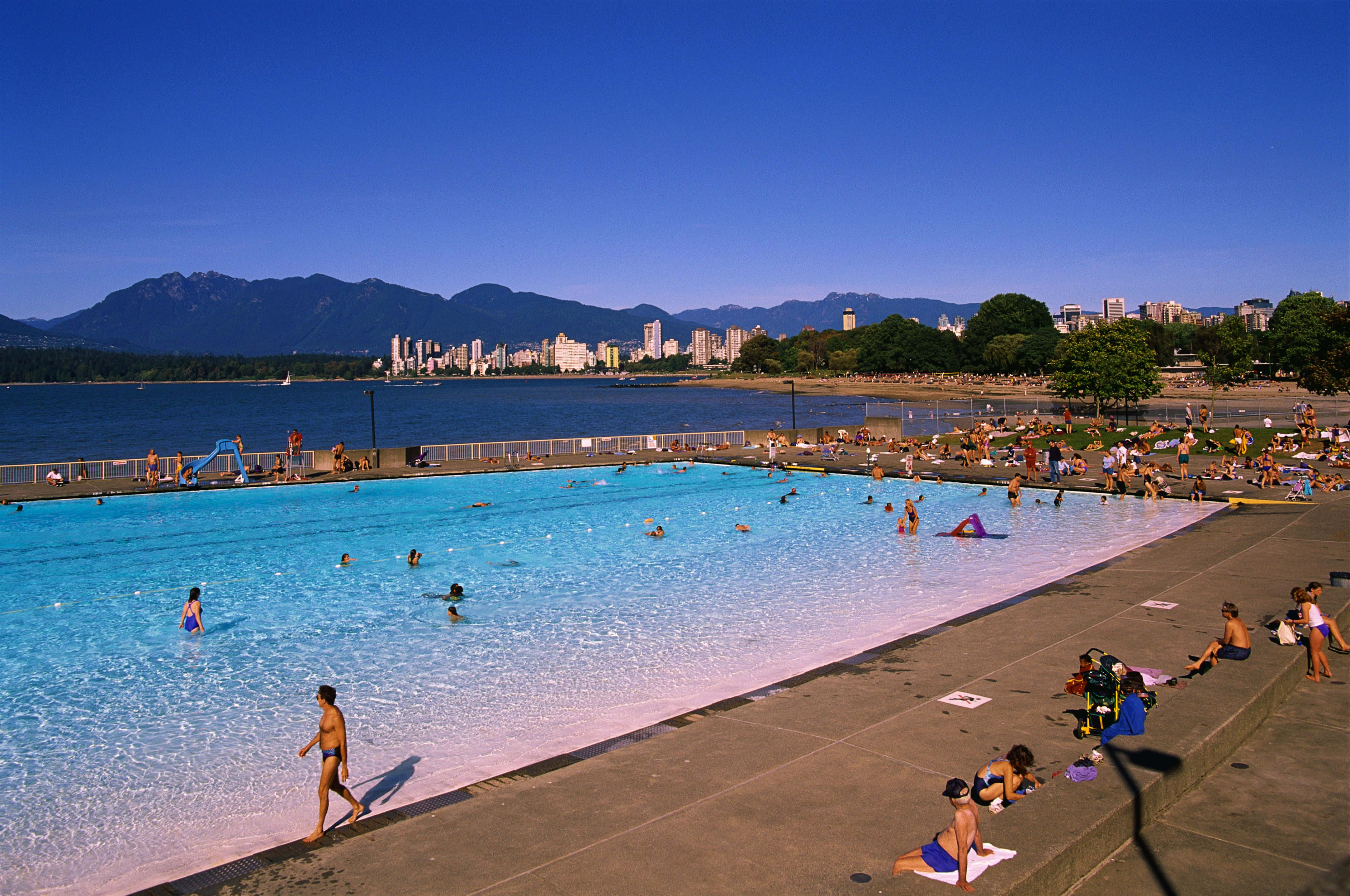Beach Swimming Pool 桌布 壁紙 — 一個熙熙攘攘的戶外游泳池，有游泳者和日光浴者，在晴朗的藍天下，遠處有山脈和城市天際線。