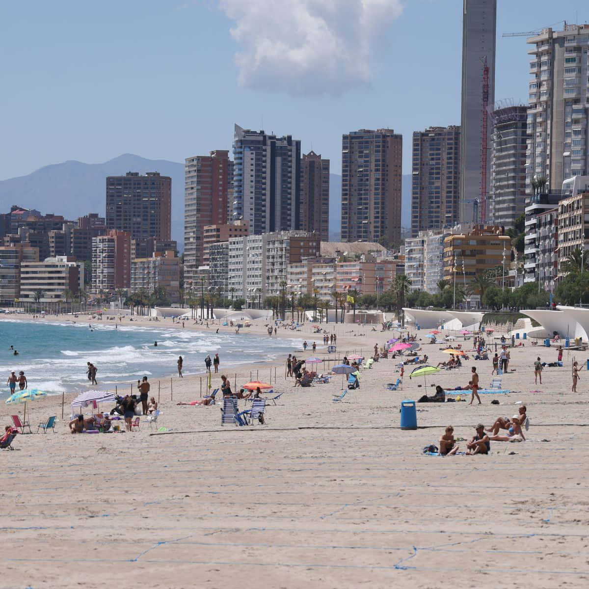 Spanish Beach 桌布 壁紙 — 一個熙熙攘攘的海灘，擠滿了享受日光浴的人們和色彩繽紛的遮陽傘，背景是明亮的藍天、現代高樓大廈和遠處的山脈。