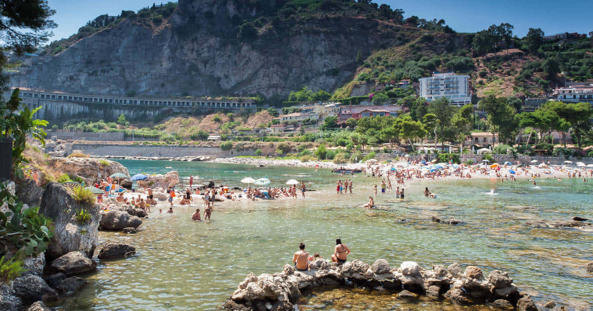 Italian Island 桌布 壁紙 — 一個熱鬧、陽光普照的海灘，清澈碧綠的海水，岩石海岸，四周環繞著翠綠的山丘和建築物。