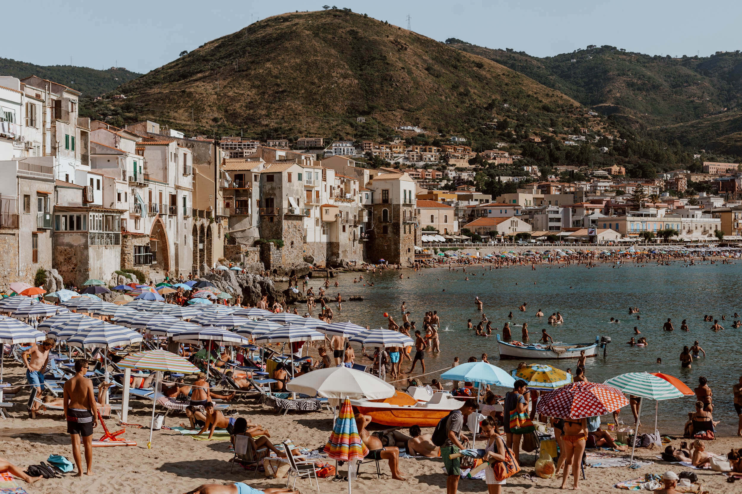 Italian Beach 桌布 壁紙 — 一個熱鬧的地中海海灘，擠滿了在彩色遮陽傘下享受日光浴的人們，背景是白色建築和山丘。