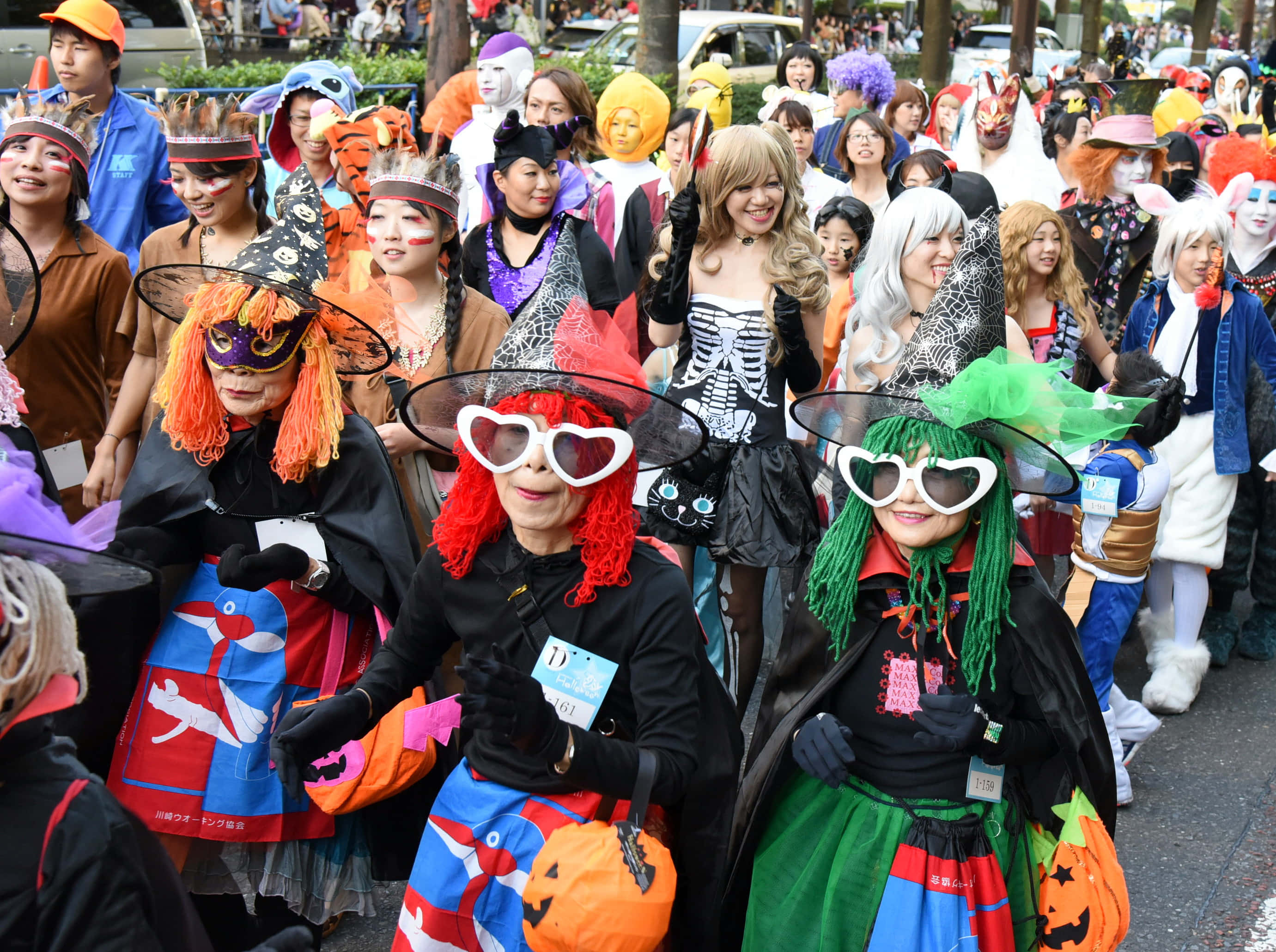 Halloween Parades 桌布 壁紙 — 一個熱鬧的萬聖節巡遊，人們穿著色彩繽紛的服裝，在繁忙的街道上微笑並擺姿勢拍照。