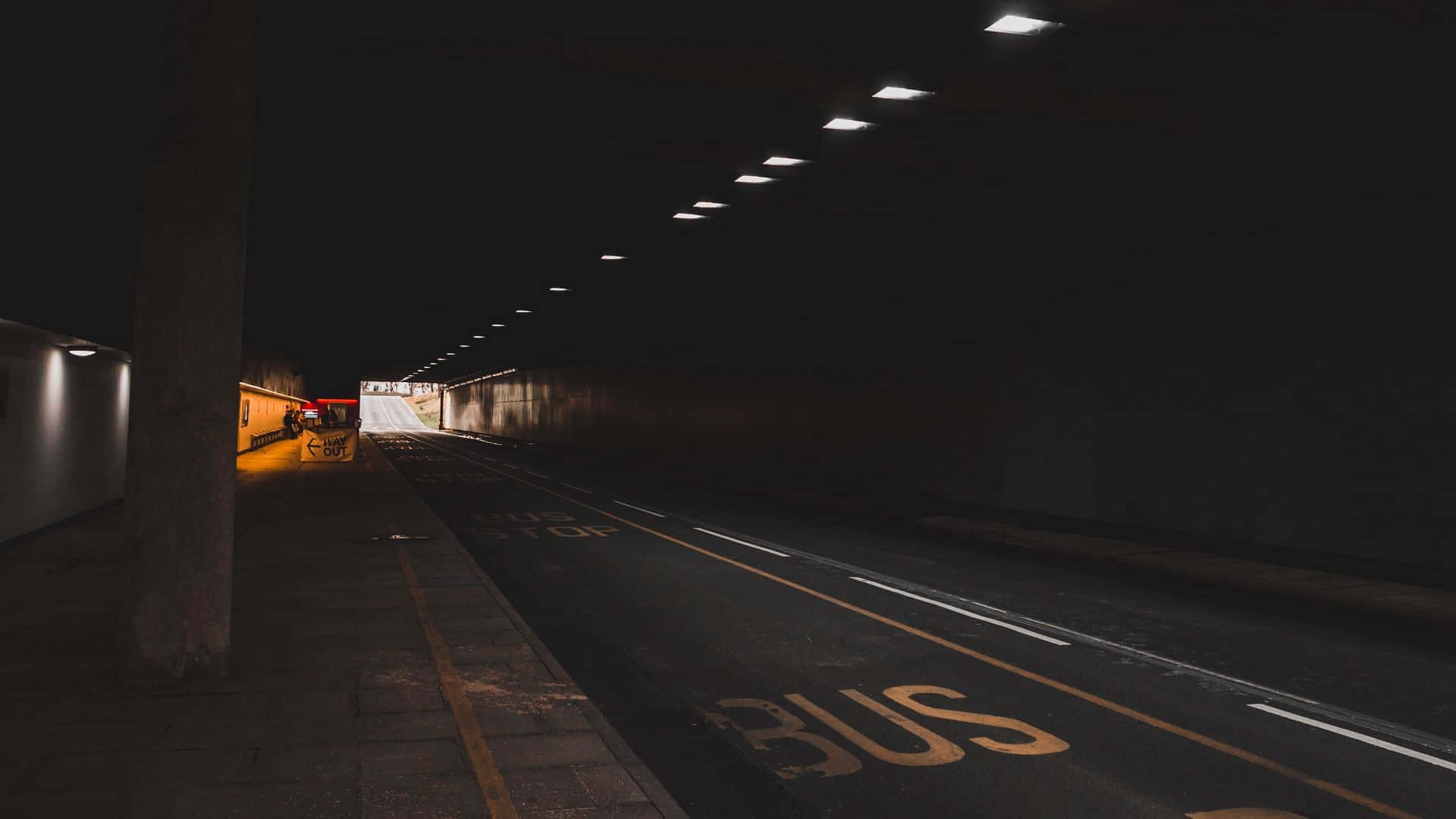 Dark Underground 桌布 壁紙 — 一個燈光昏暗的城市隧道，頭頂燈光在道路上標記的巴士站上投下長長的陰影。
