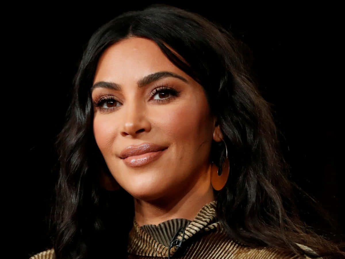 Kim Kardashian 桌布 壁紙 — 一個特寫肖像，一位擁有深色波浪長髮和大圈耳環的女性，背景為黑色。
